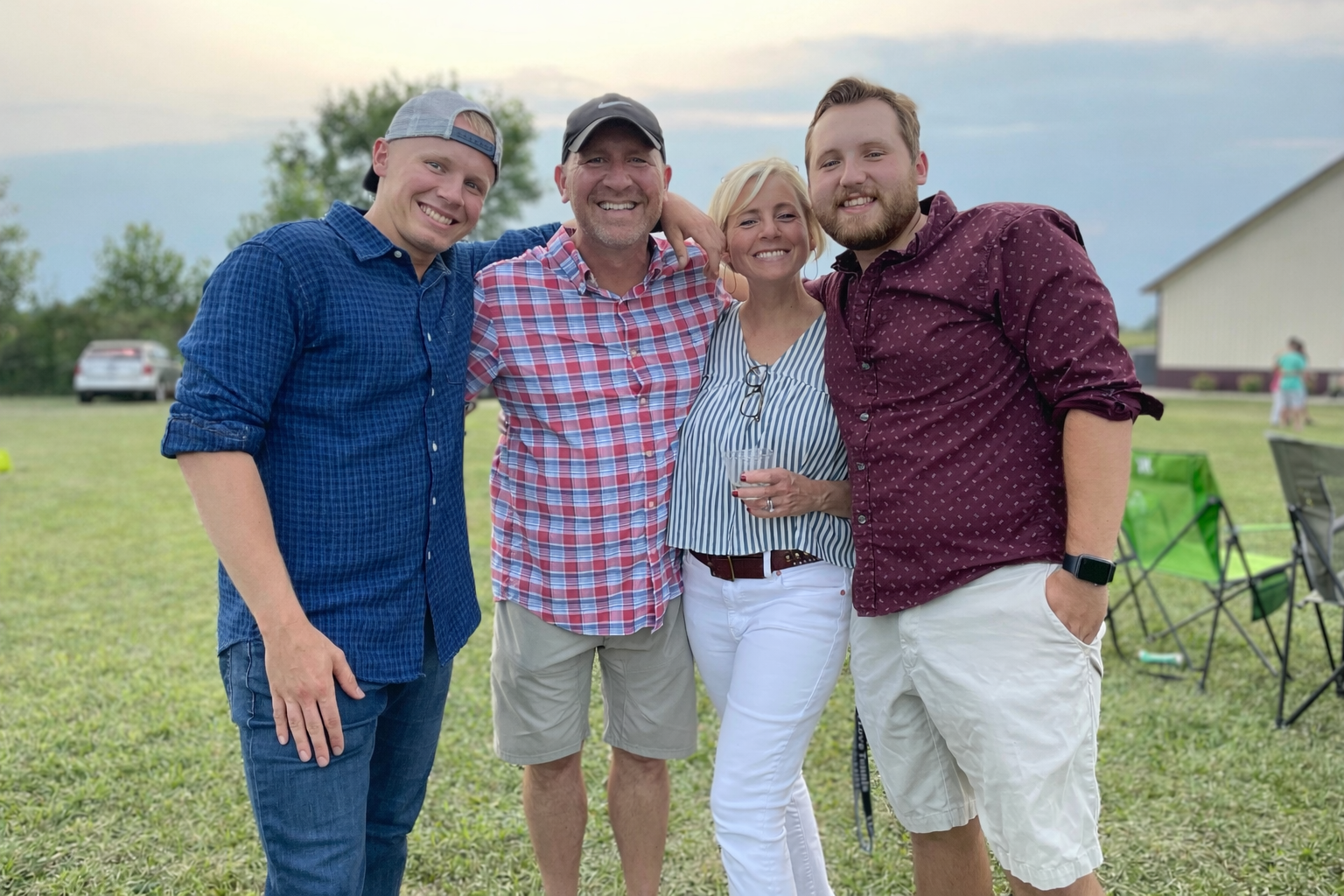 A group of four adults standing outdoors on a grassy field, smiling and posing for the camera. They include two men and one woman in the center, and a man on the right. There are chairs and other people in the background, with a building and trees under an overcast sky.