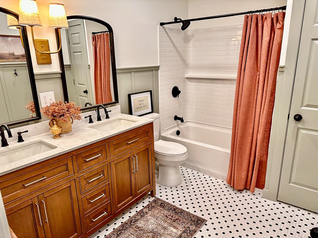 A bathroom with a double vanity featuring a white marble countertop, black fixtures, and wooden cabinets. There are two mirrors above the sinks, a framed quote that says "Today is a good day," and a vase with pink flowers on the counter. The room inc