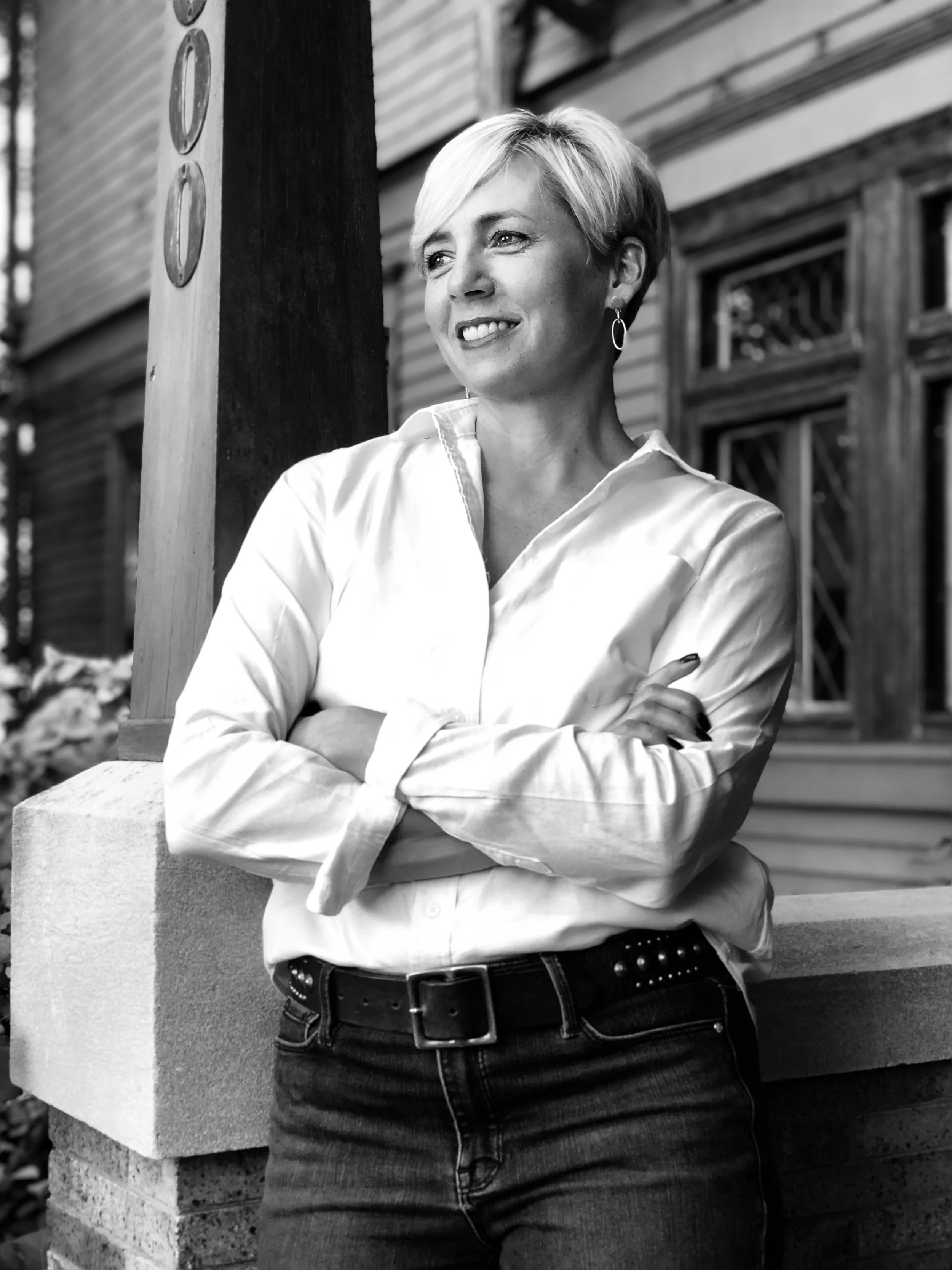 Staci Slavin, Head Designer and Owner, a woman with short blonde hair, wearing a white button-up shirt and dark jeans, standing outside in front of a house with wooden siding, smiling and looking to her right with crossed arms.