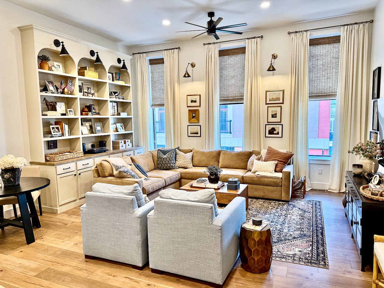 Living room with beige sectional sofa, light gray armchairs, a wooden coffee table, a black side table, a white built-in bookshelf, and a black console table. There are four tall windows with cream curtains and brown woven shades, gold wall sconces, 