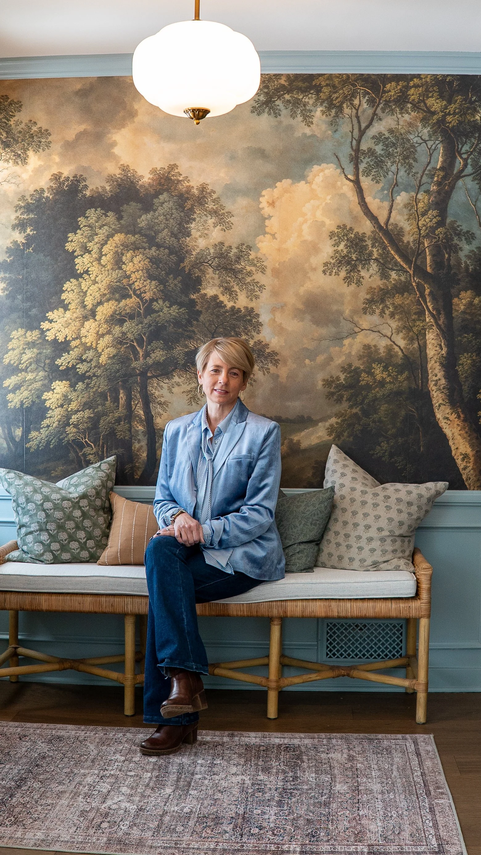 Staci Slavin, Head Designer and Owner, A woman sitting on a wooden bench in front of a large mural of trees and clouds, with decorative pillows behind her, in a room with a hanging ceiling light.