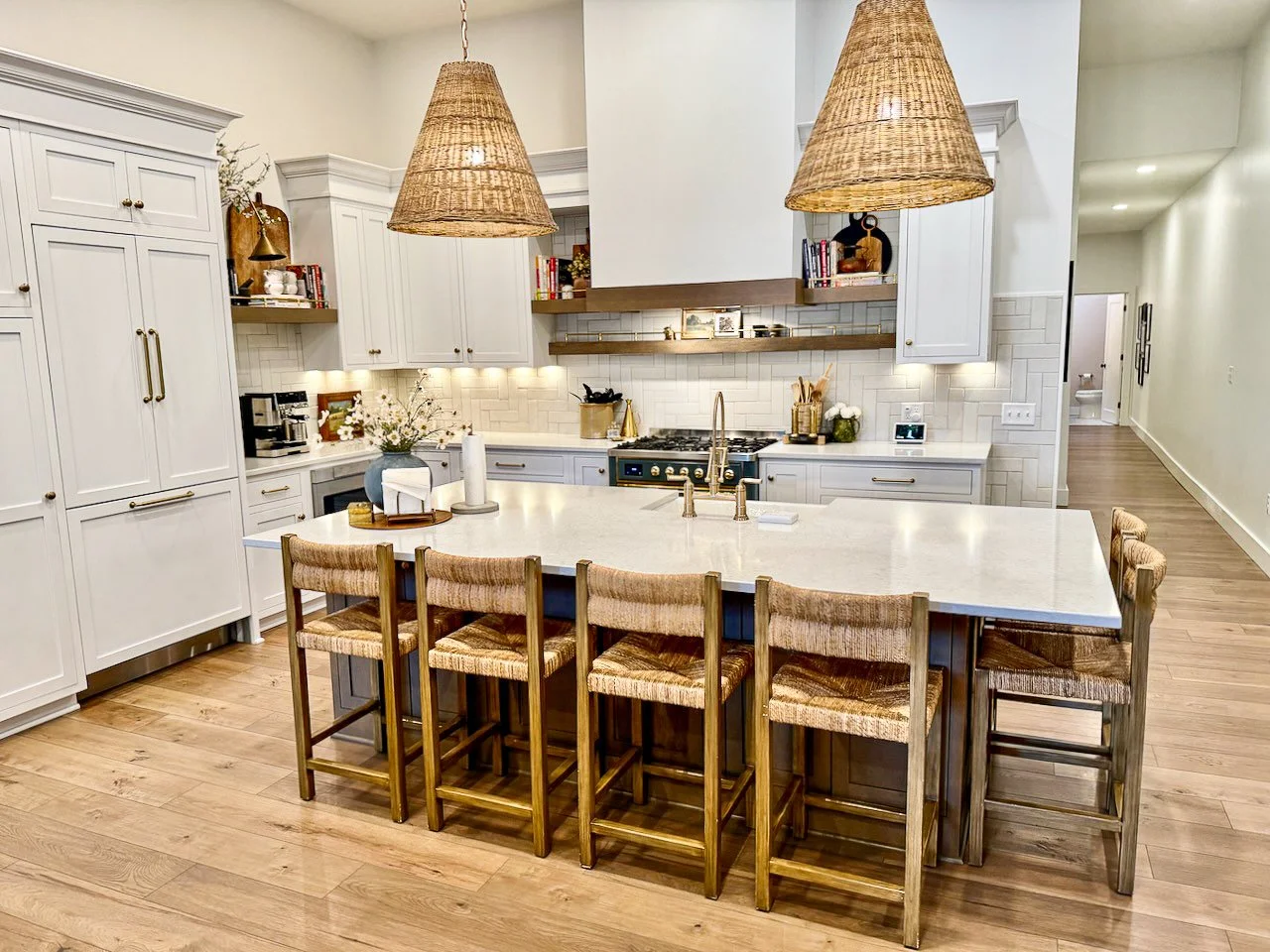 Modern kitchen with white cabinetry, a large island with six wooden and woven chairs, and two pendant lights made of woven material hanging above the island. The background features a stove, open shelves with books and decor, and a light-colored wood
