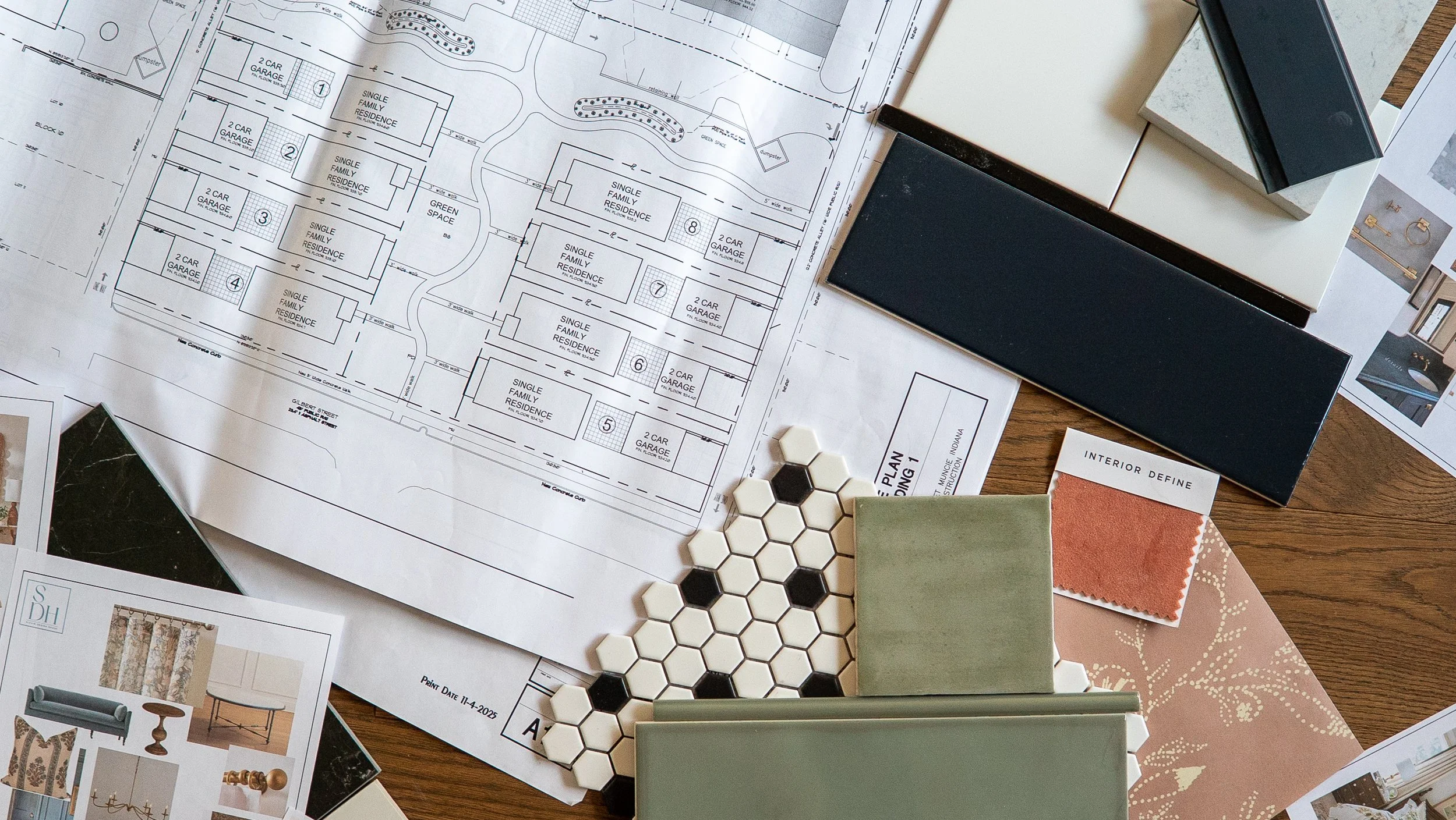 A top-down view of an architectural blueprint spread on a wooden table, surrounded by design materials including color swatches, tile samples, fabric swatches, and printed interior design images.