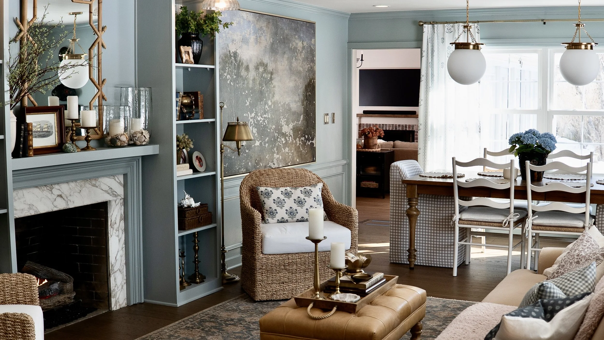 Living room with blue walls, fireplace with marble surround, blue shelving with decor, wicker chair with patterned pillow, wooden dining table with chairs, large window with white curtains, blue flowers on table, and various decorative items.