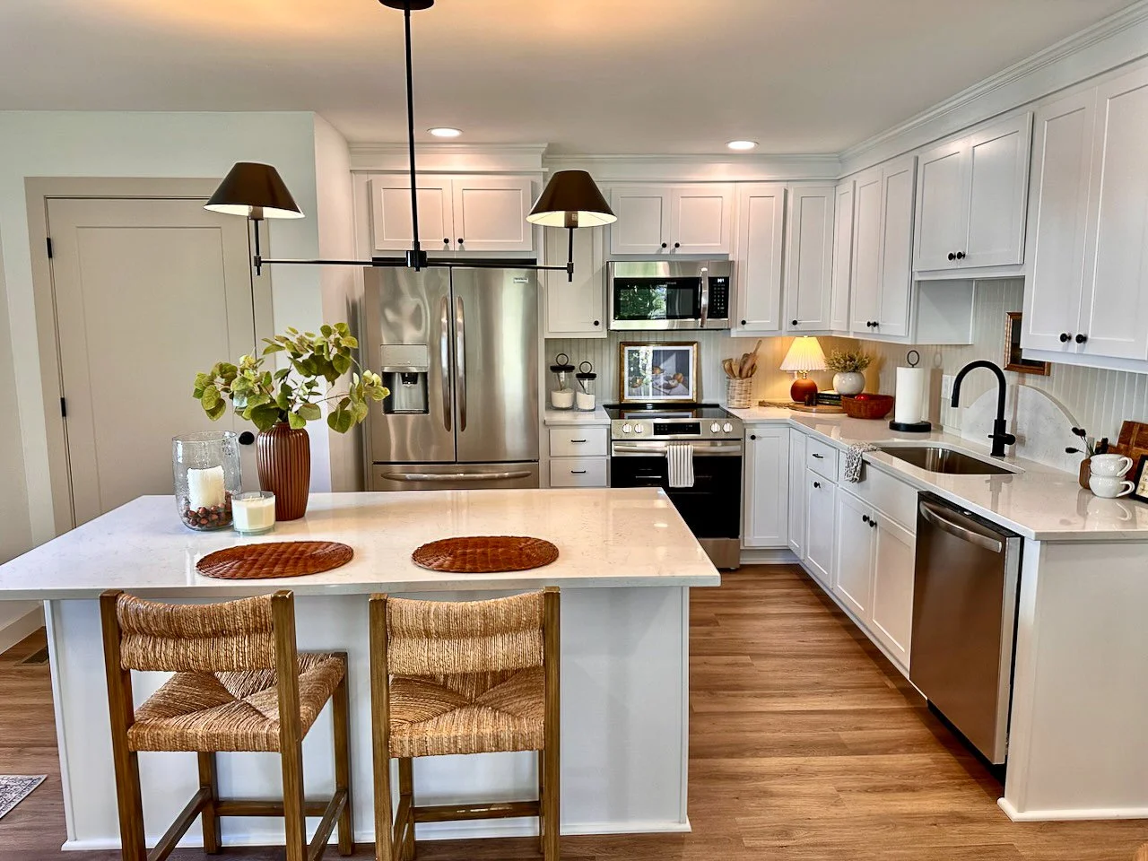 Bright kitchen with white cabinets, stainless steel refrigerator, microwave, and dishwasher. An island with two wicker barstools, a vase with greenery, candles, and woven placemats. Counter with decorative items, sink, and black faucet. Wooden floori