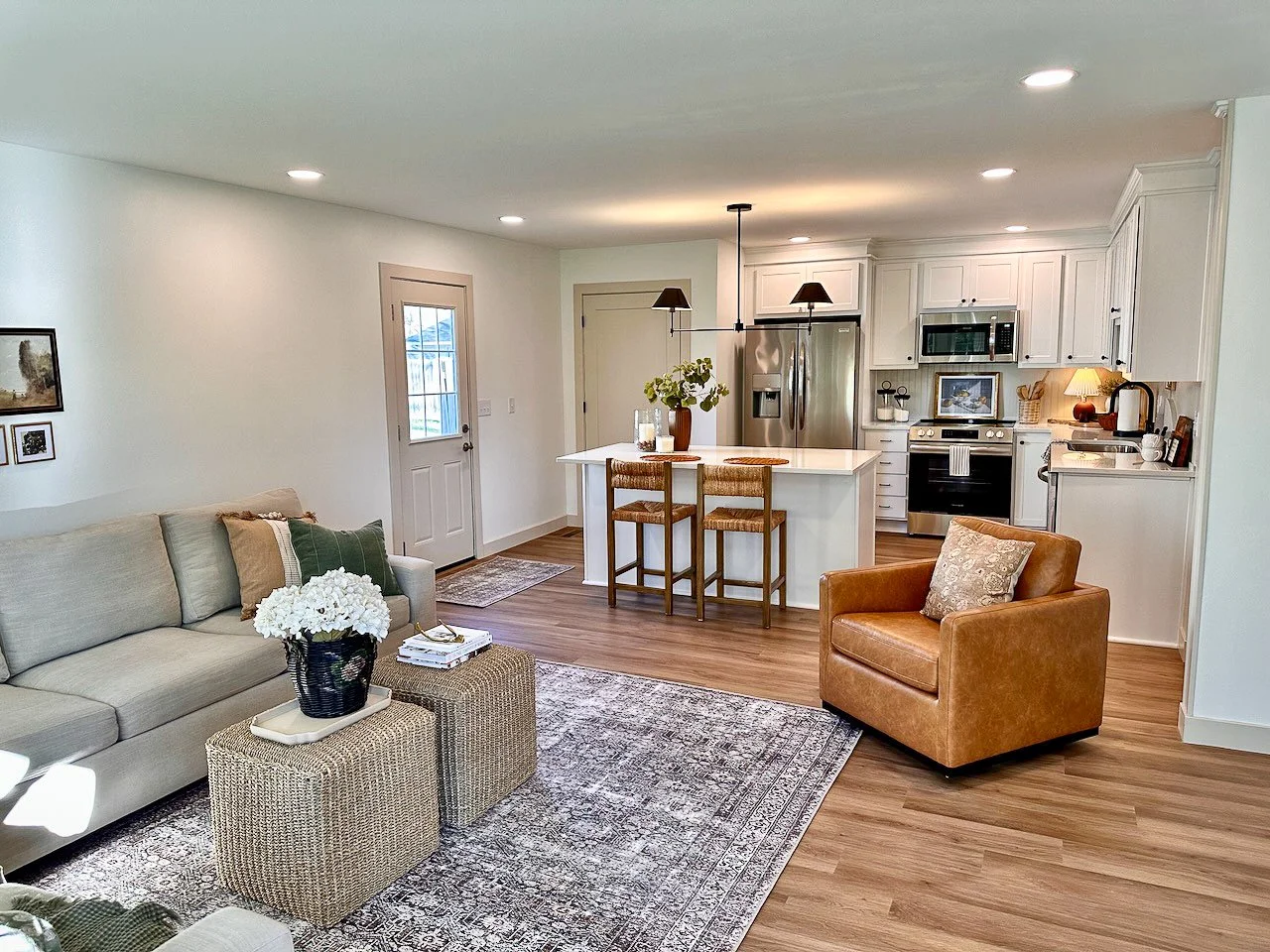 Open-concept living room and kitchen with white cabinets, stainless steel appliances, and hardwood floors. Furnished with a beige sofa, brown armchair, patterned rug, and decorative items.