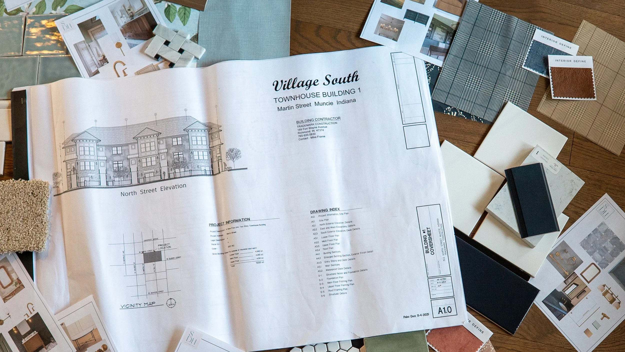 Architectural plans and interior design samples spread on a wooden table, including a large blueprint for a townhouse building, sample fabrics, paint swatches, and color tiles.