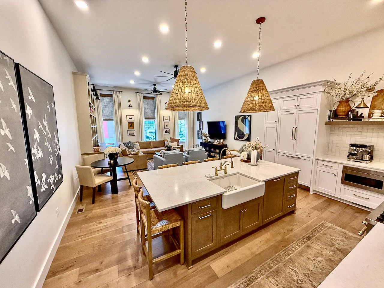 Open-concept living room and kitchen with white cabinetry, wooden flooring, and a central island with a white sink. Decor includes framed art, a coffee station, hanging rattan pendant lights, and cozy seating area with beige and gray sofas.