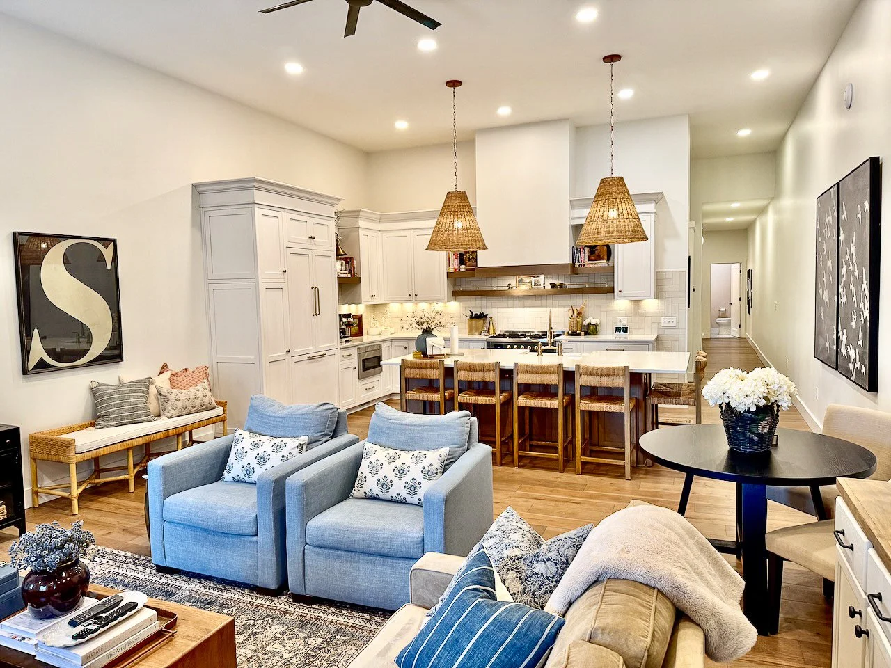 Open-concept living room and kitchen with white cabinetry, pendant lights, gray armchairs, and a round black dining table with a flower vase.