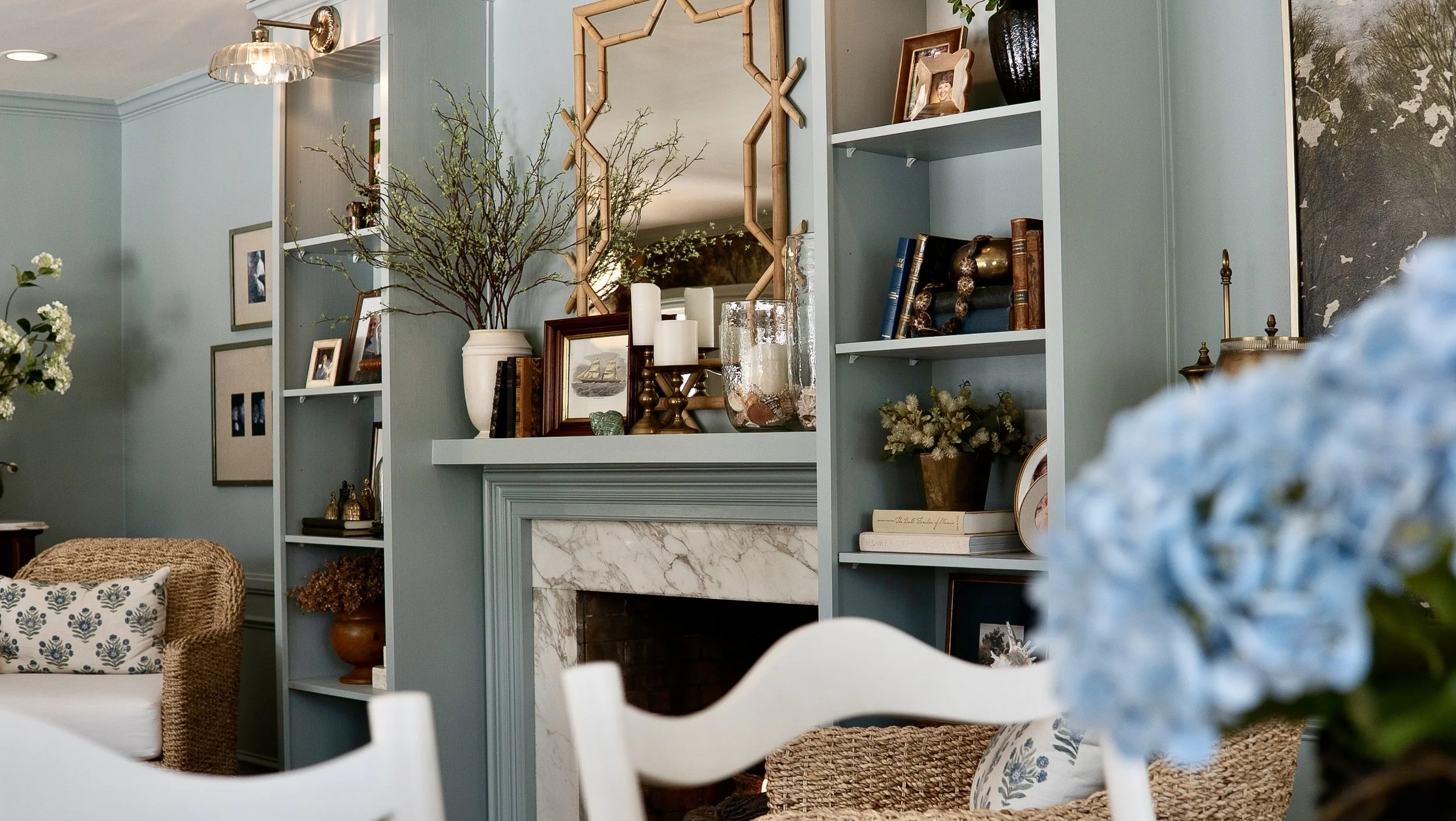 Interior of a living room with built-in blue shelves above a marble fireplace, decorated with books, framed photos, vases, candles, and plants, with wicker furniture and floral cushions, and a large blue hydrangea flower in the foreground.