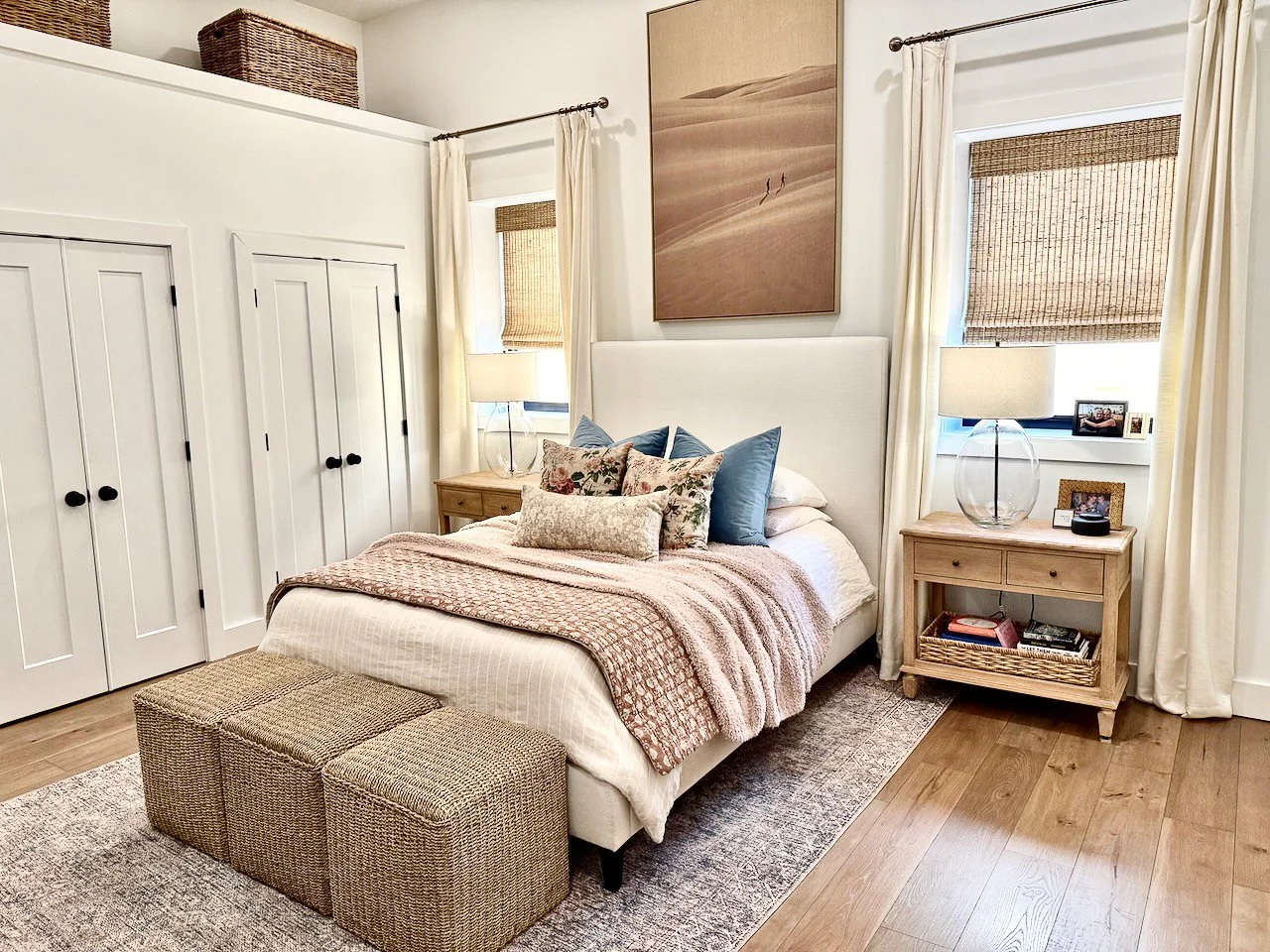 A cozy bedroom with a white bed, multiple decorative pillows, two wooden nightstands with lamps, and a large desert landscape painting above the bed. There are two windows with bamboo shades and white curtains, a carpet under the bed, and woven baske