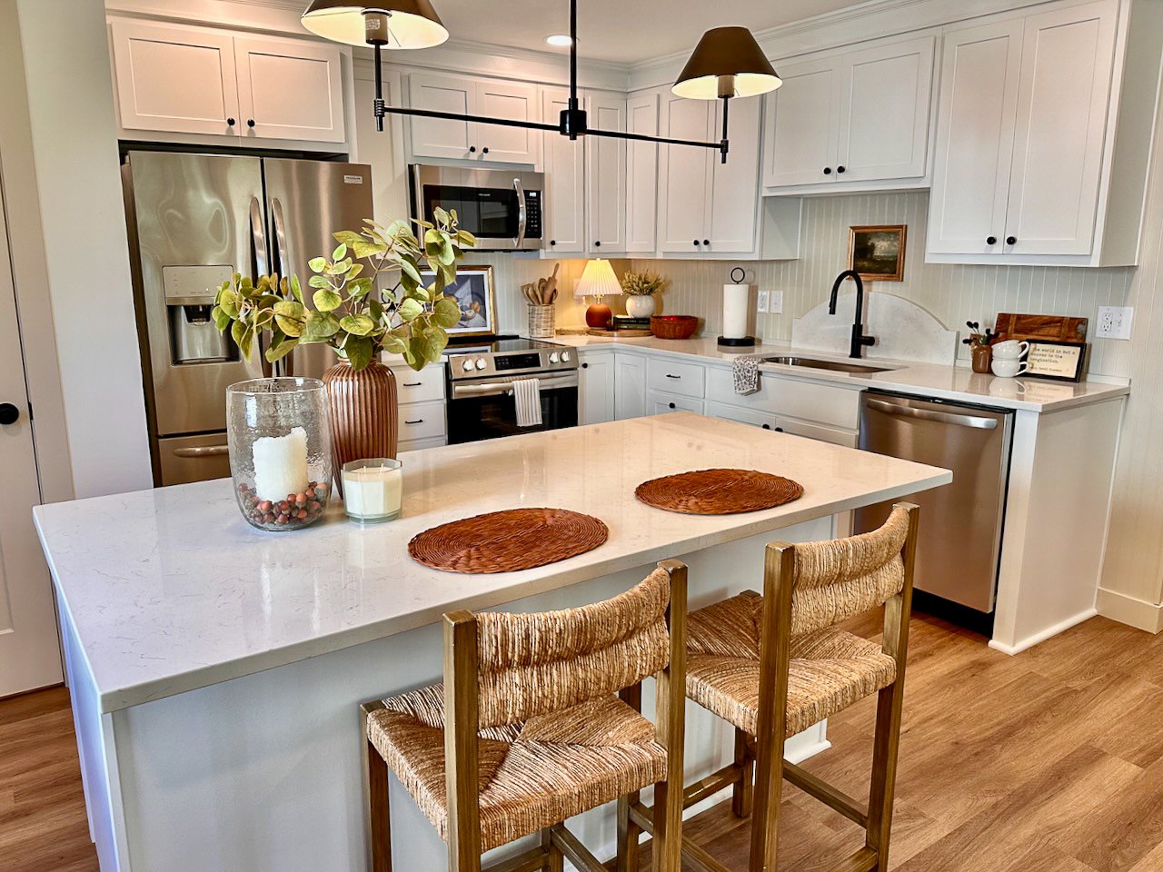 A modern kitchen with white cabinets, stainless steel appliances, a kitchen island with a white marble countertop, and wooden bar stools. Decor includes a vase with green leaves, candles, and various small decorative items.