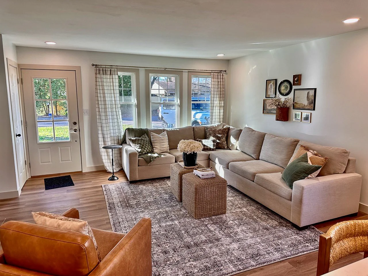 Cozy living room with beige sectional sofa, wooden coffee table, armchair, and large window with checkered curtains, decorated with framed art and a potted plant.