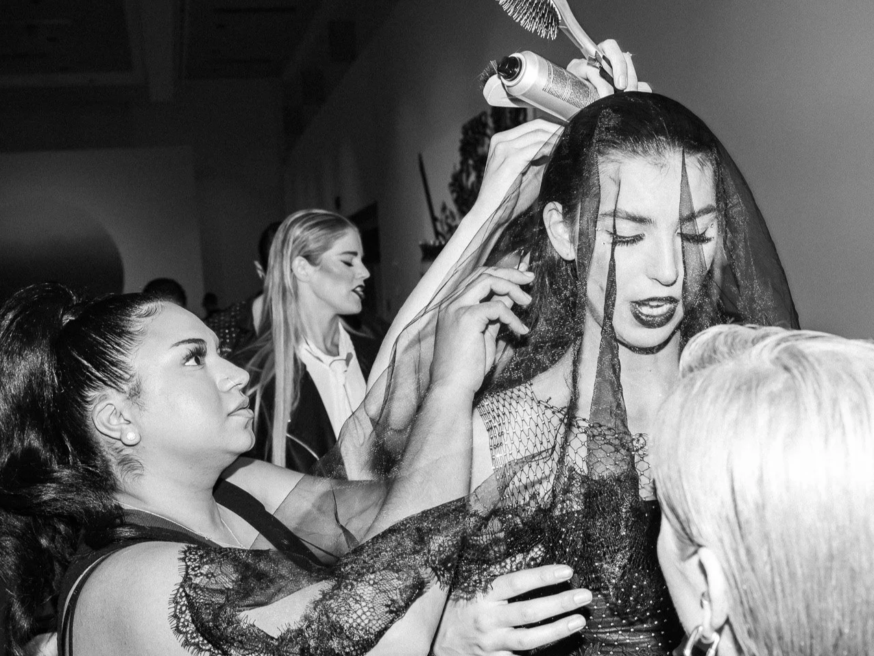 A woman with dark lipstick and lace clothing getting her hair styled with spray by a hairstylist at a themed event, with other women in the background.