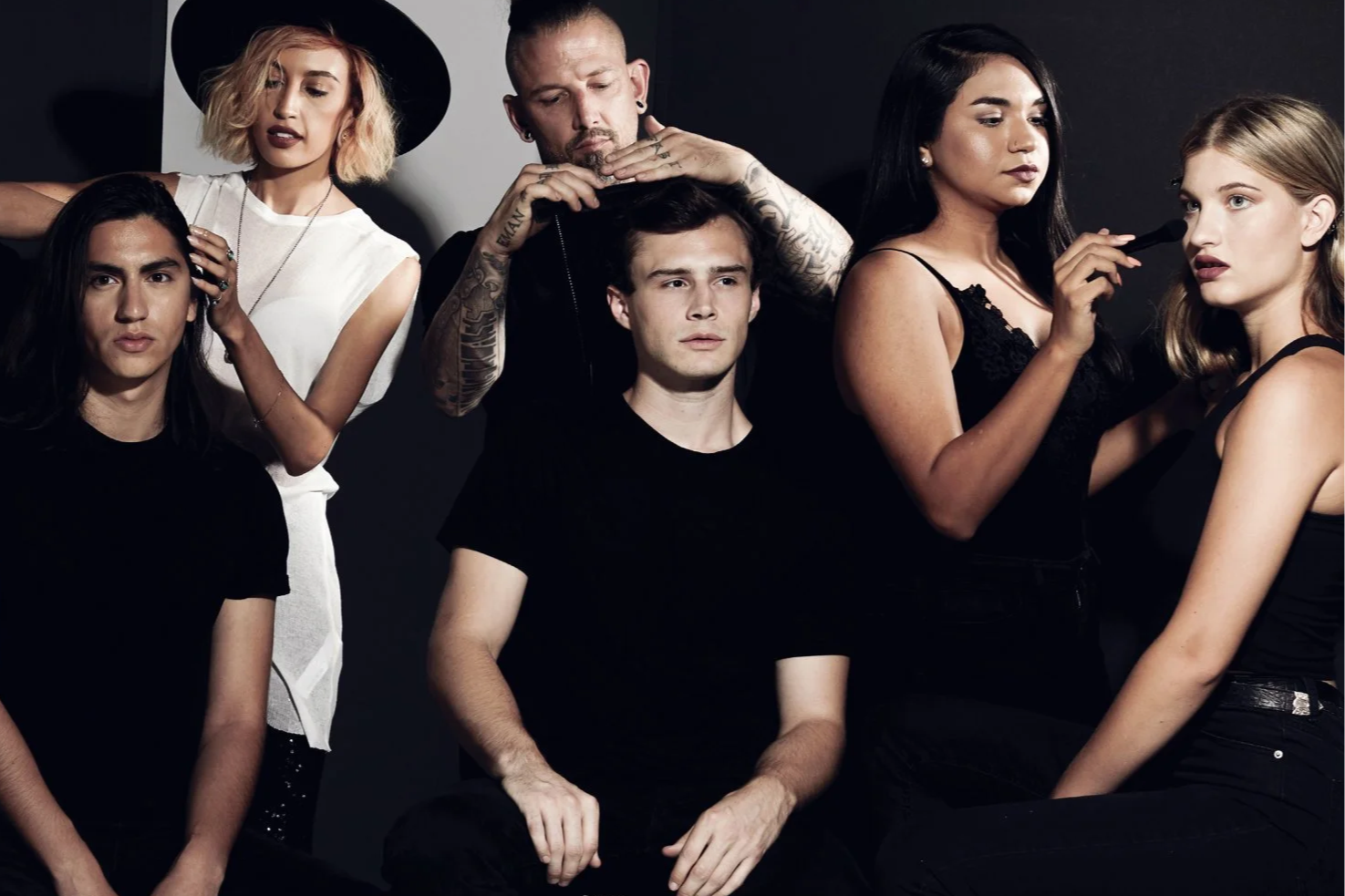 A group of six young adults, three women and three men, with various hair colors and styles, in a dark room with a black background. One woman is applying makeup to another woman, while the others are either looking at the camera or looking away, with a serious or focused expression.