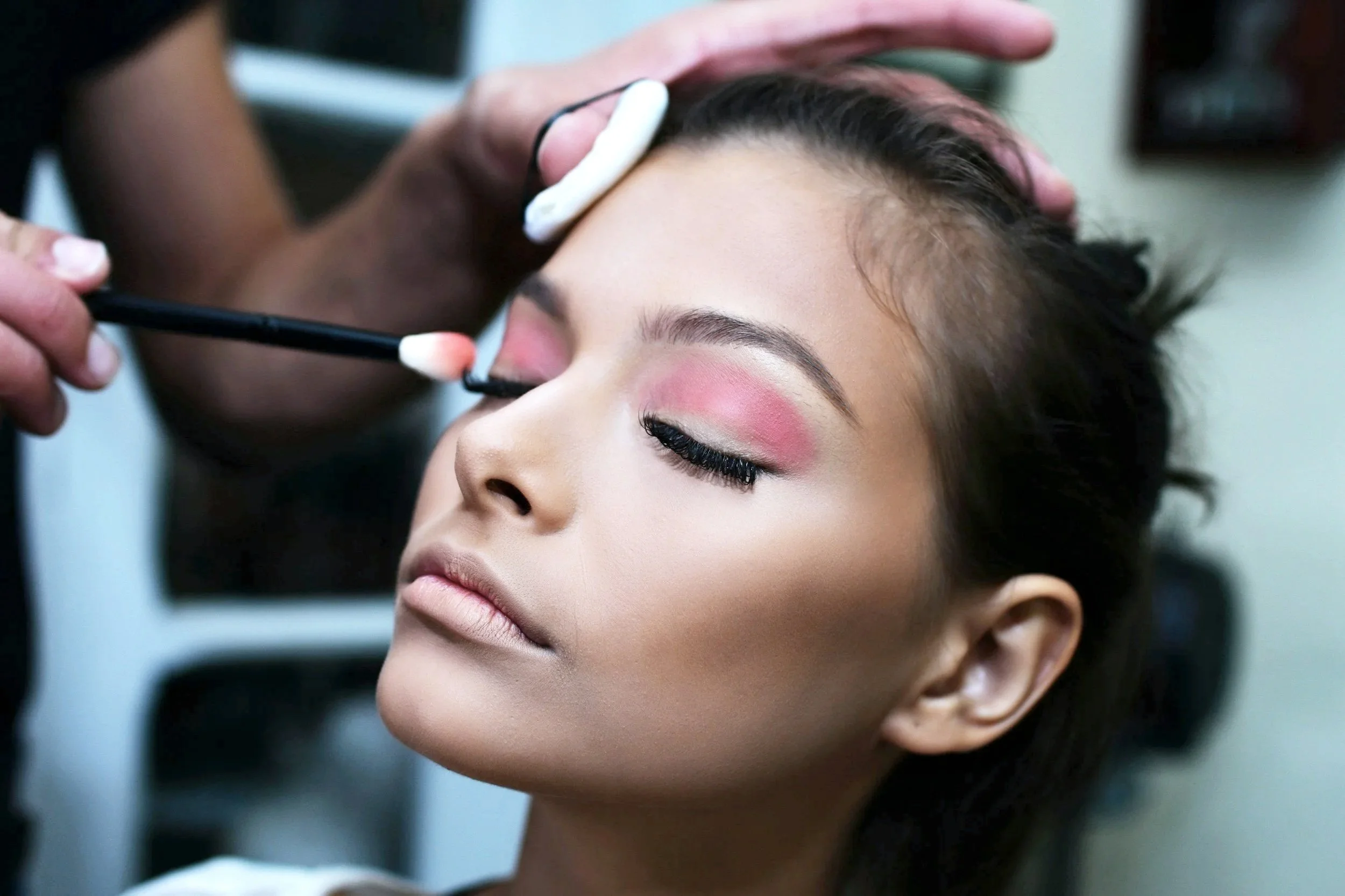 A woman receiving makeup application, with eyes closed, as makeup artist applies eye makeup with brushes.