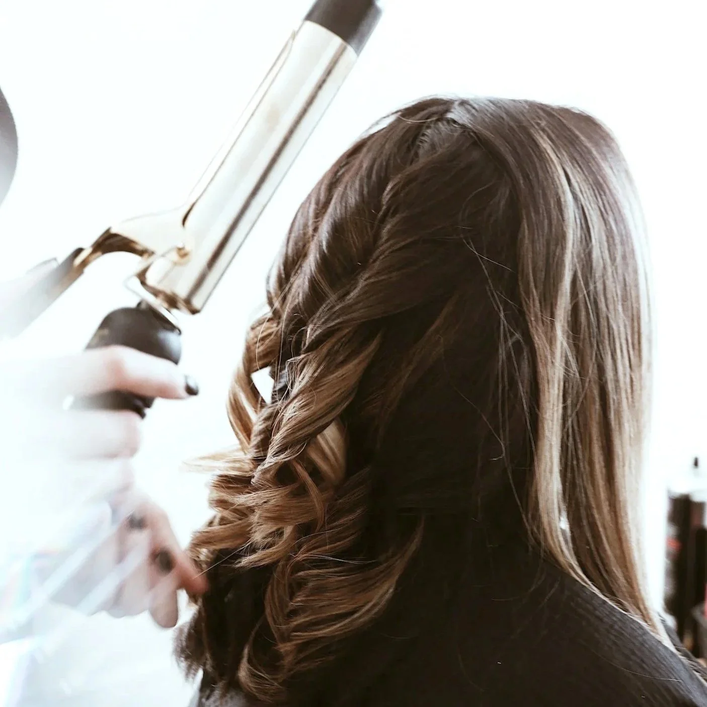 A woman with brown hair styled in loose curls is getting her hair dried with a hairdryer.