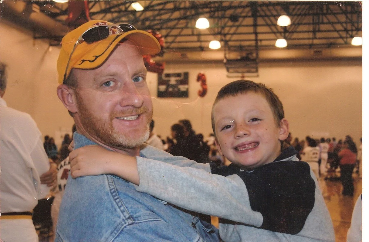 A man wearing a yellow cap with sunglasses on it, and a light blue denim jacket, holding a smiling young boy with freckles, in an indoor gymnasium filled with people.