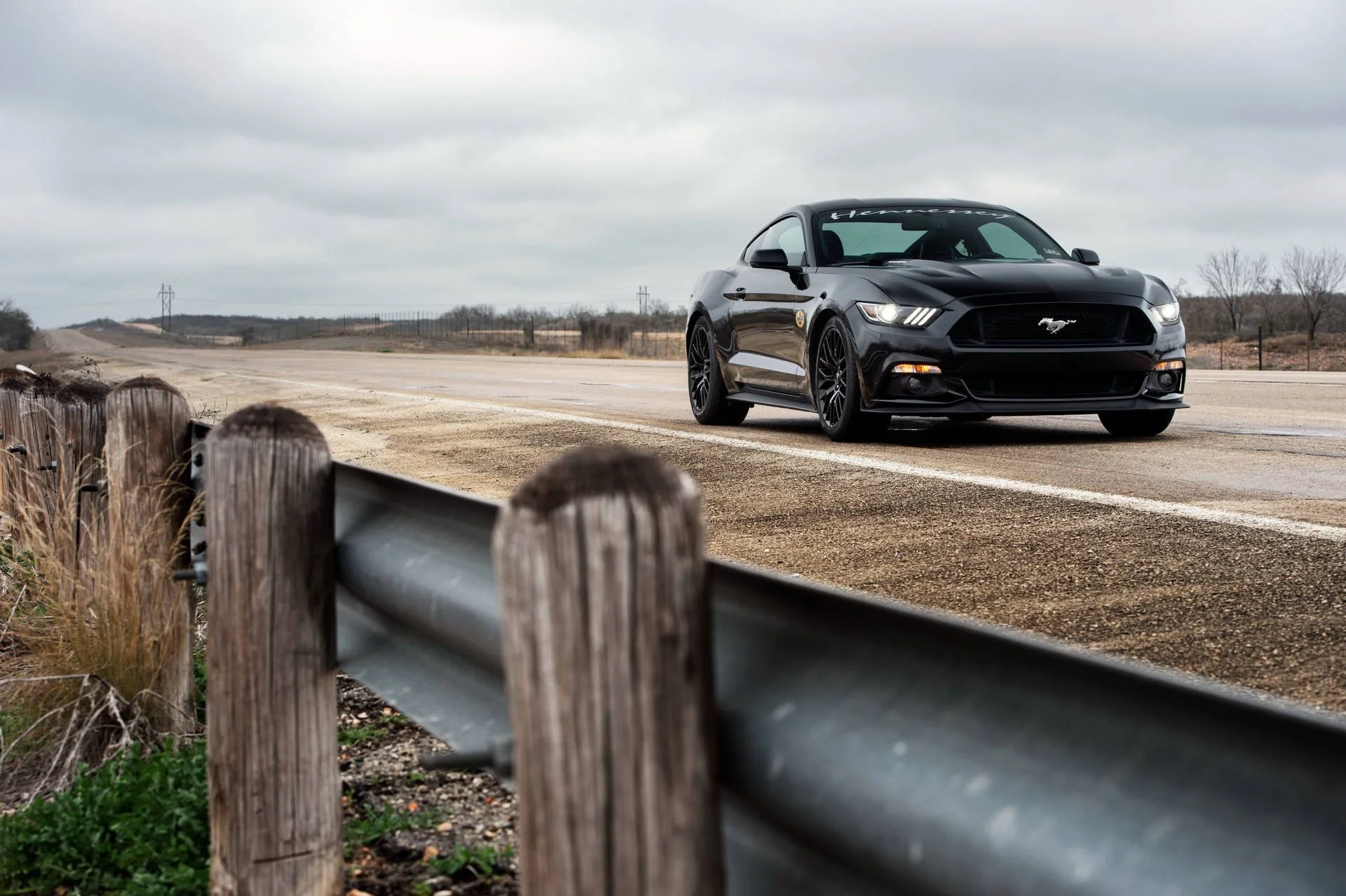 A black Ford Mustang on a rural two-lane road under cloudy skies, with a wooden guardrail in the foreground and open land in the background.