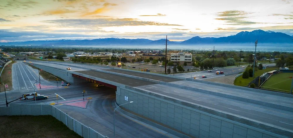 View of a highway overpass and roads at sunrise, mountains in the background, with some cars and buildings in the distance.