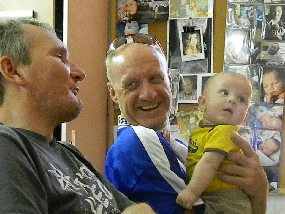 Two adult males, one holding a baby, are sitting close together indoors near a wall decorated with numerous photographs and pictures.