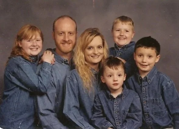 A family portrait of six people, including a man, woman, and four children, all wearing denim shirts, smiling against a gray backdrop.