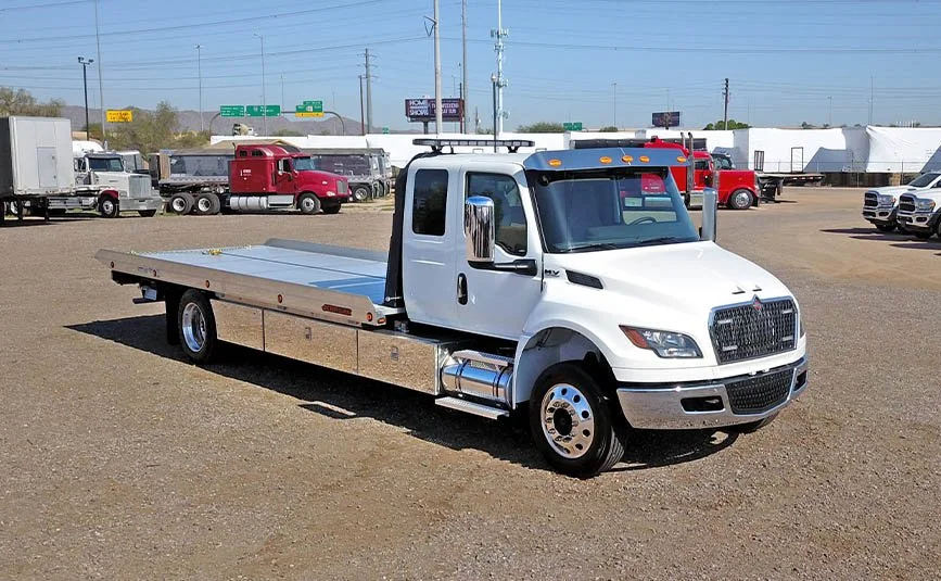 White flatbed tow truck parked on a gravel lot, with other trucks and vehicles in the background.