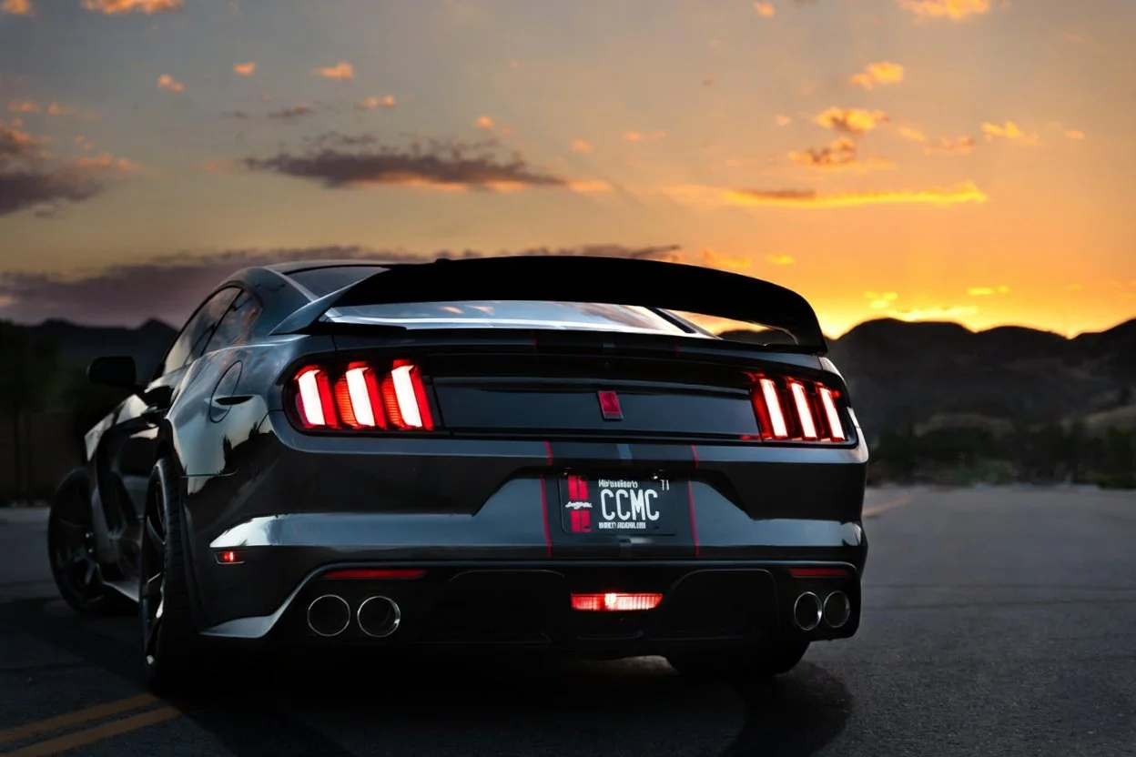 A black sports car with a spoiler and quad exhaust pipes parked on a road during sunset with mountains in the background.