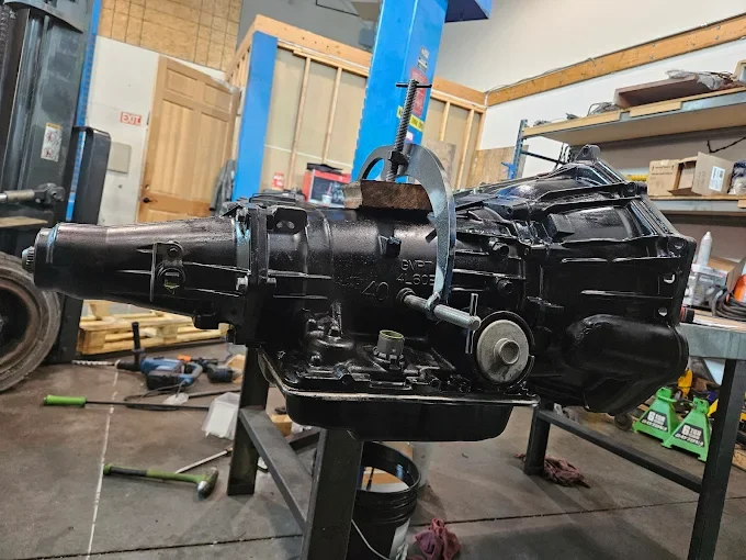 A car transmission on a workbench in a garage or workshop with tools and shelves in the background.