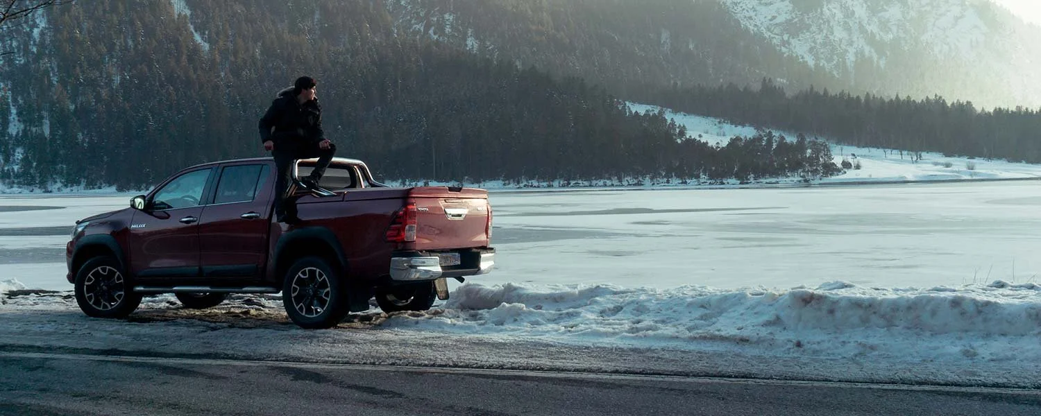 A person standing on the edge of a red pickup truck parked beside a snowy, icy lake with mountains and forest in the background.