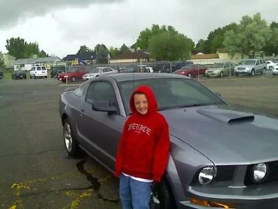 A young boy wearing a red hoodie and jeans standing next to a dark gray sports car in a parking lot on a cloudy day.