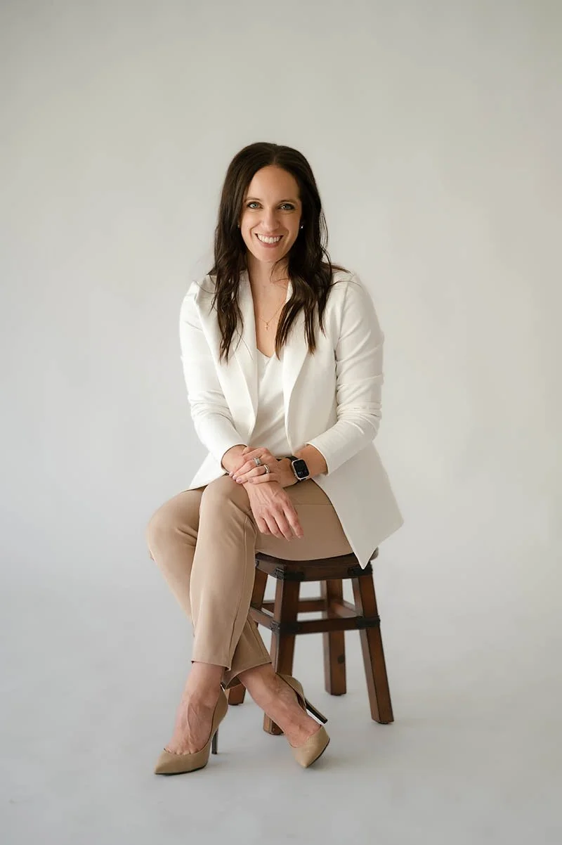 A woman with dark brown hair, wearing a white blazer, beige pants, and beige heels, sitting on a wooden stool, smiling at the camera.