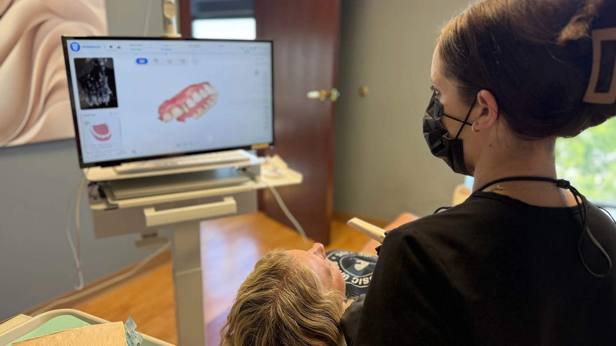 A woman is examining a seated patient with blonde hair, in a dental care setting with a computer screen displaying images.