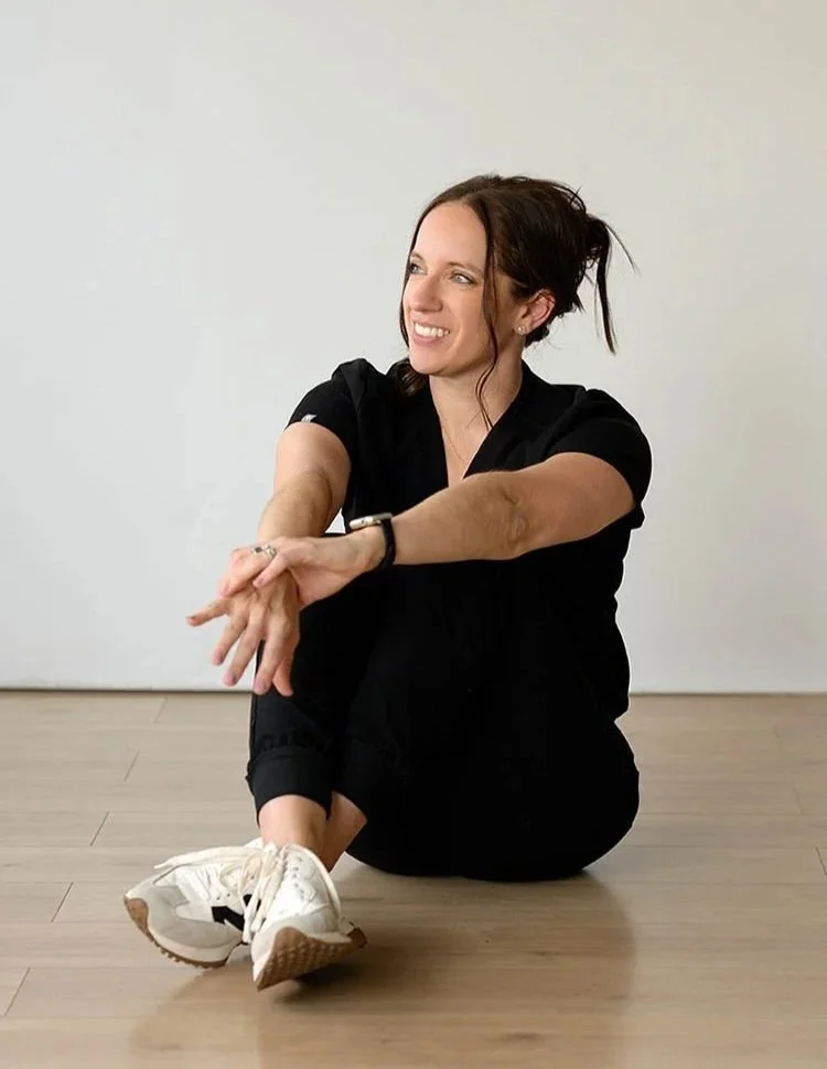 A woman sitting on the floor, dressed in black athletic wear, with a smile, in a room with light-colored walls and wooden flooring.