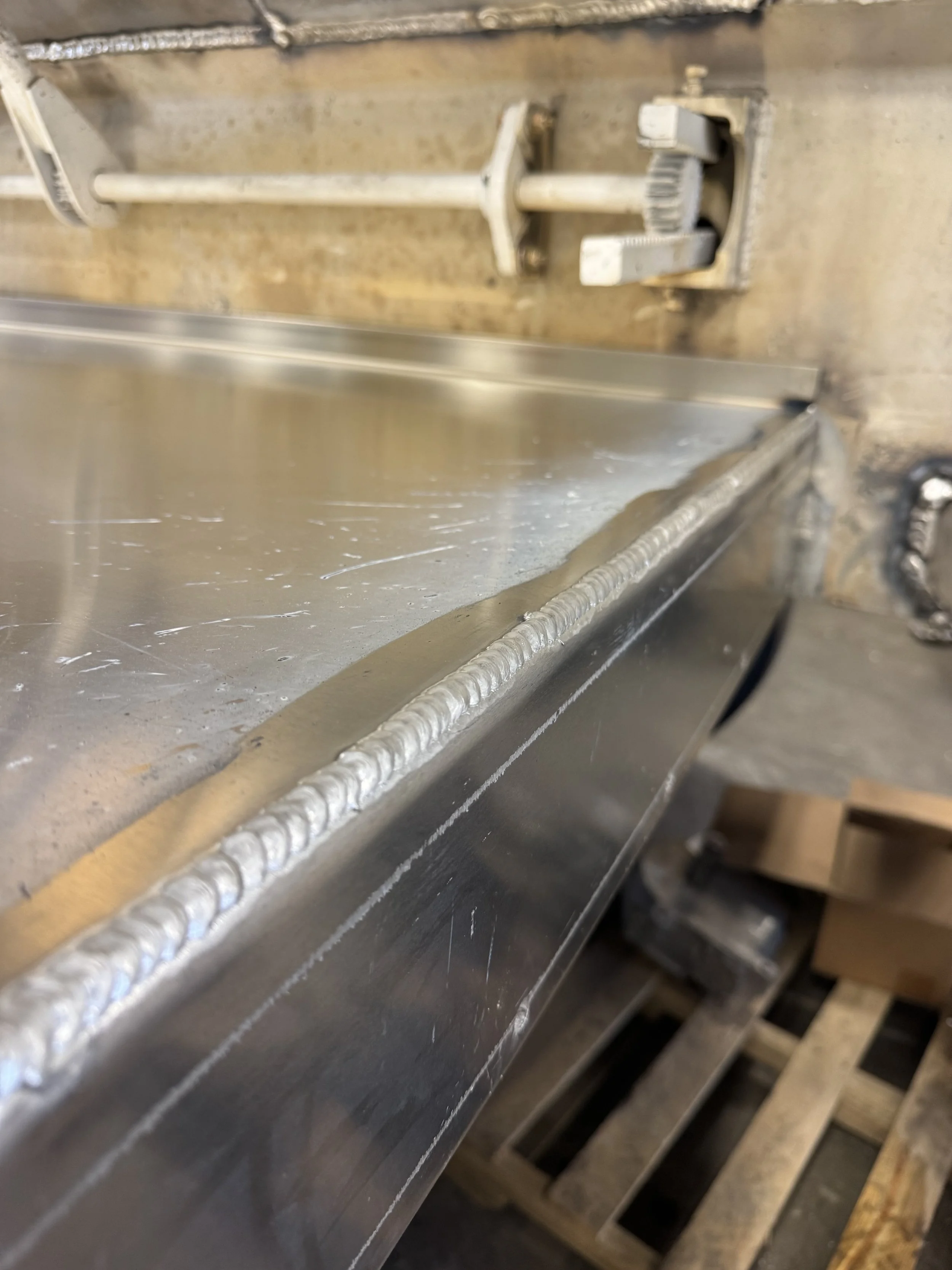 Close-up of a metal workpiece with a weld bead along the edge, in a workshop setting with tools and equipment in the background.