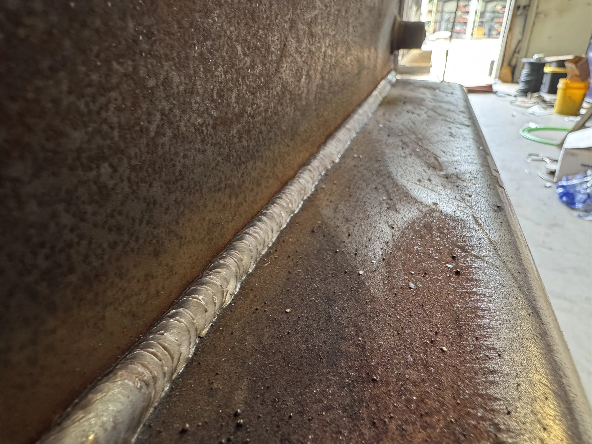 Close-up of a metal surface with a freshly welded seam running along the edge, with a workshop or construction site in the background.