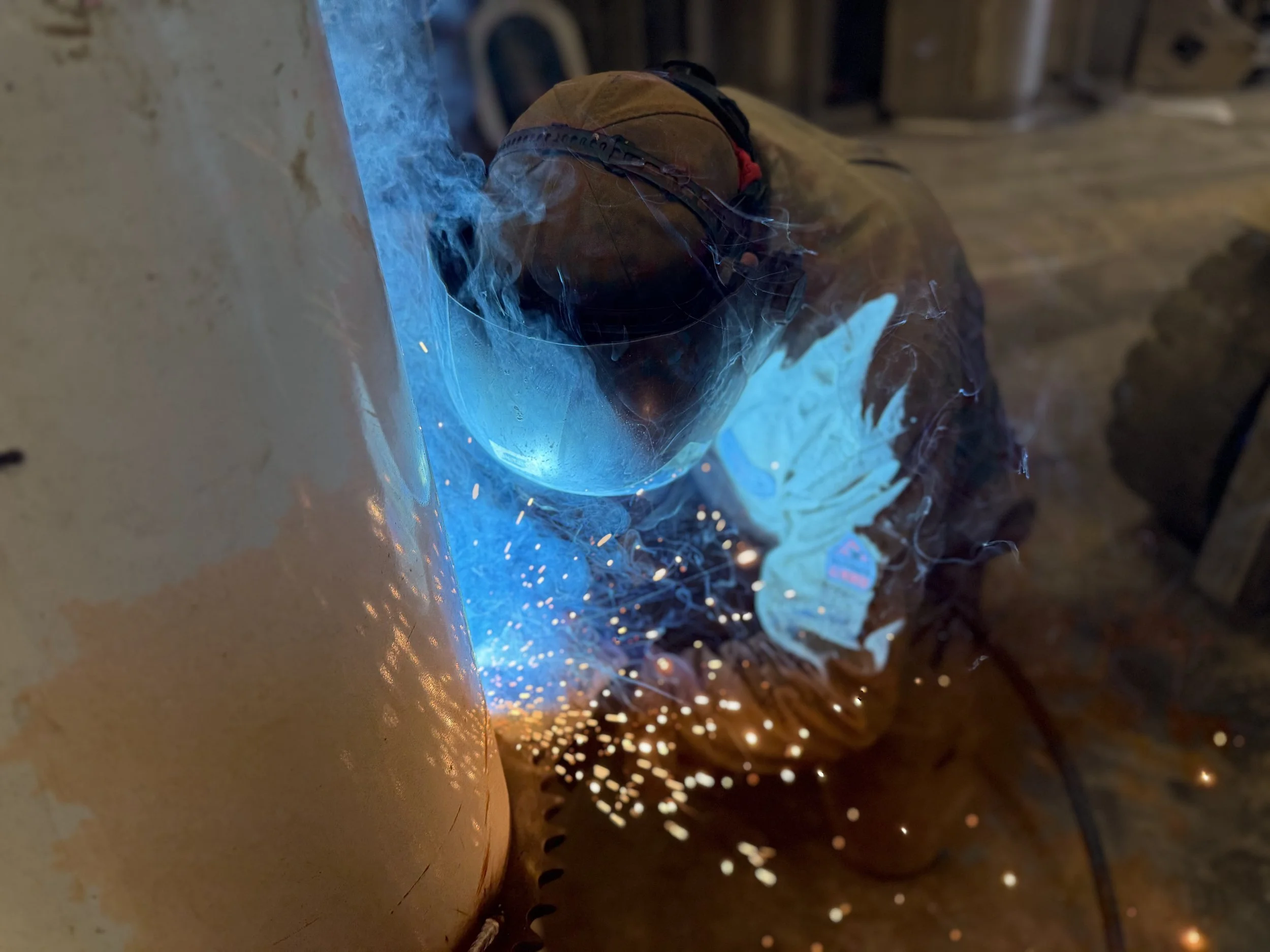 Welder wearing protective mask and gloves welding metal, sparks and bright blue arc visible.