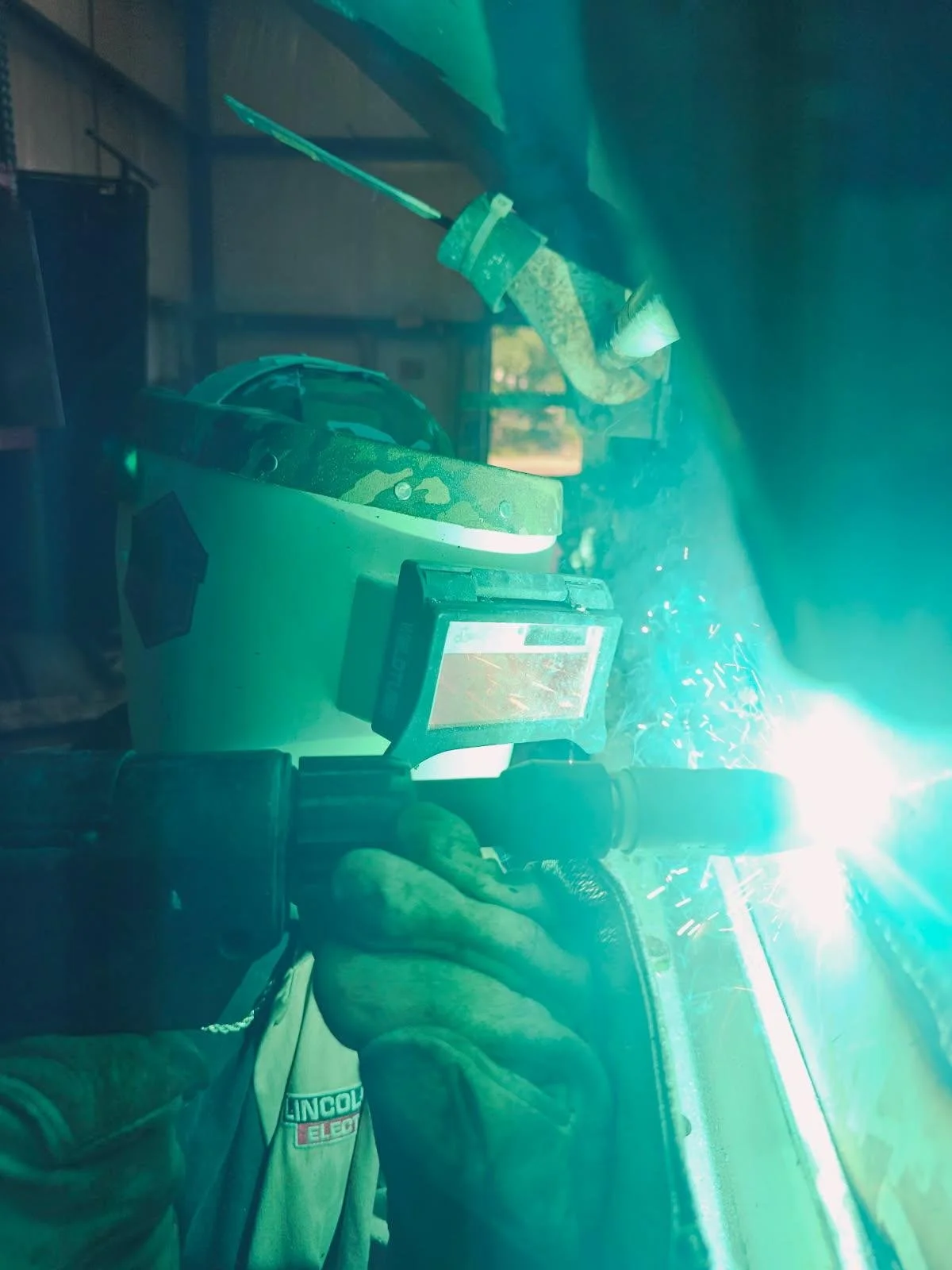Welding torch emitting bright light and sparks, welder's gloved hand holding the torch, safety helmet resting nearby, workshop with metal piping and equipment in the background.
