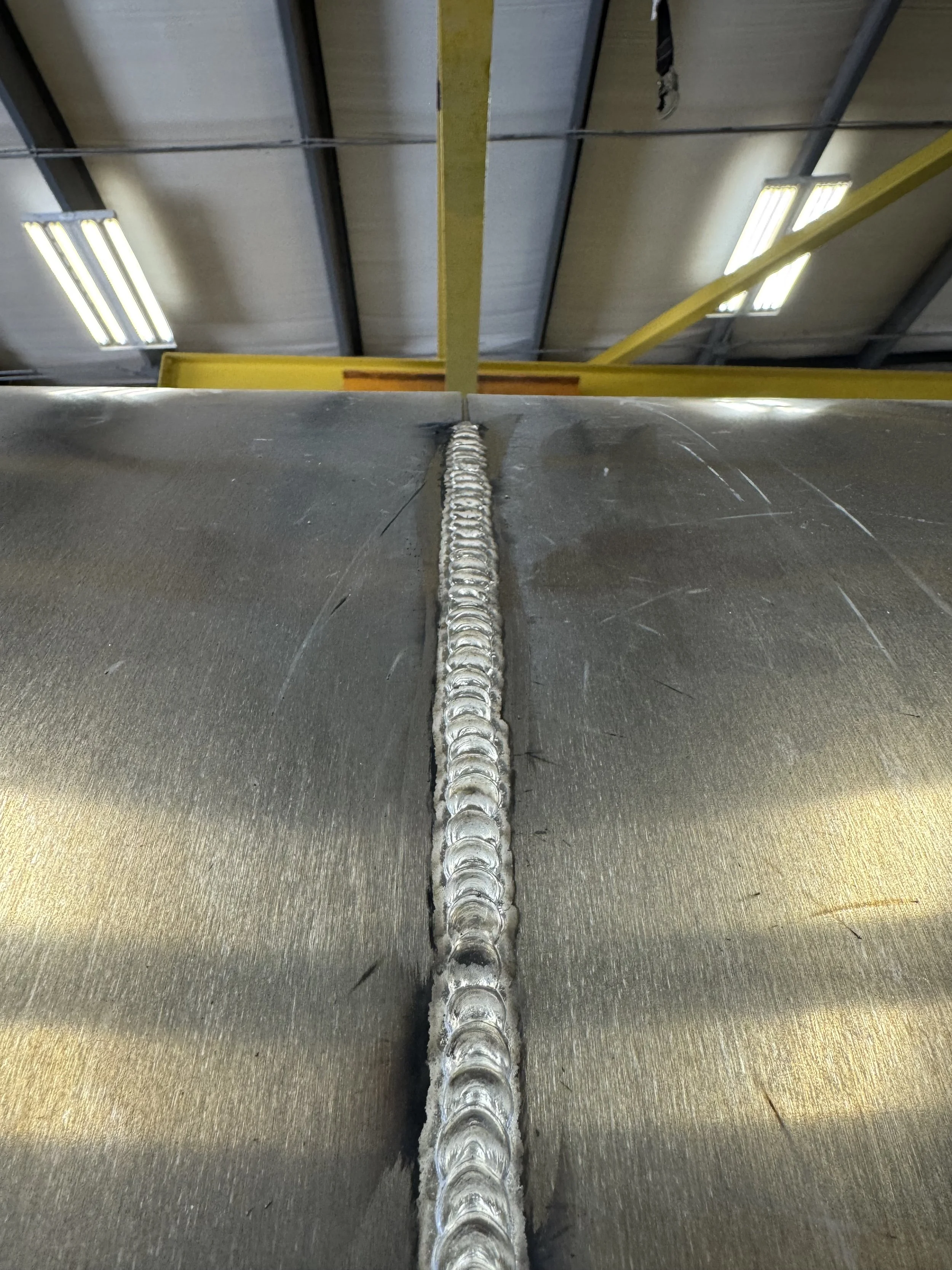 Close-up of a metal surface with a weld seam running through the center, inside an industrial or manufacturing facility with overhead lights and yellow support beams.