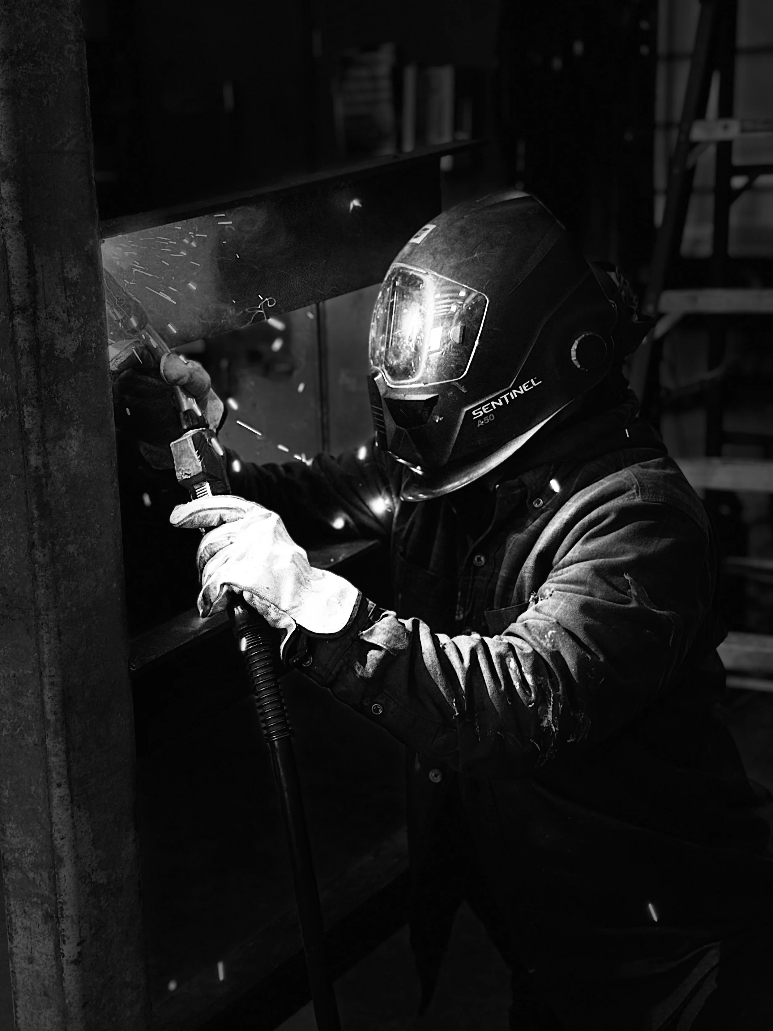 A worker welding metal in a workshop, wearing a helmet and gloves, with sparks flying as they weld a piece of metal to a larger structure in black and white.