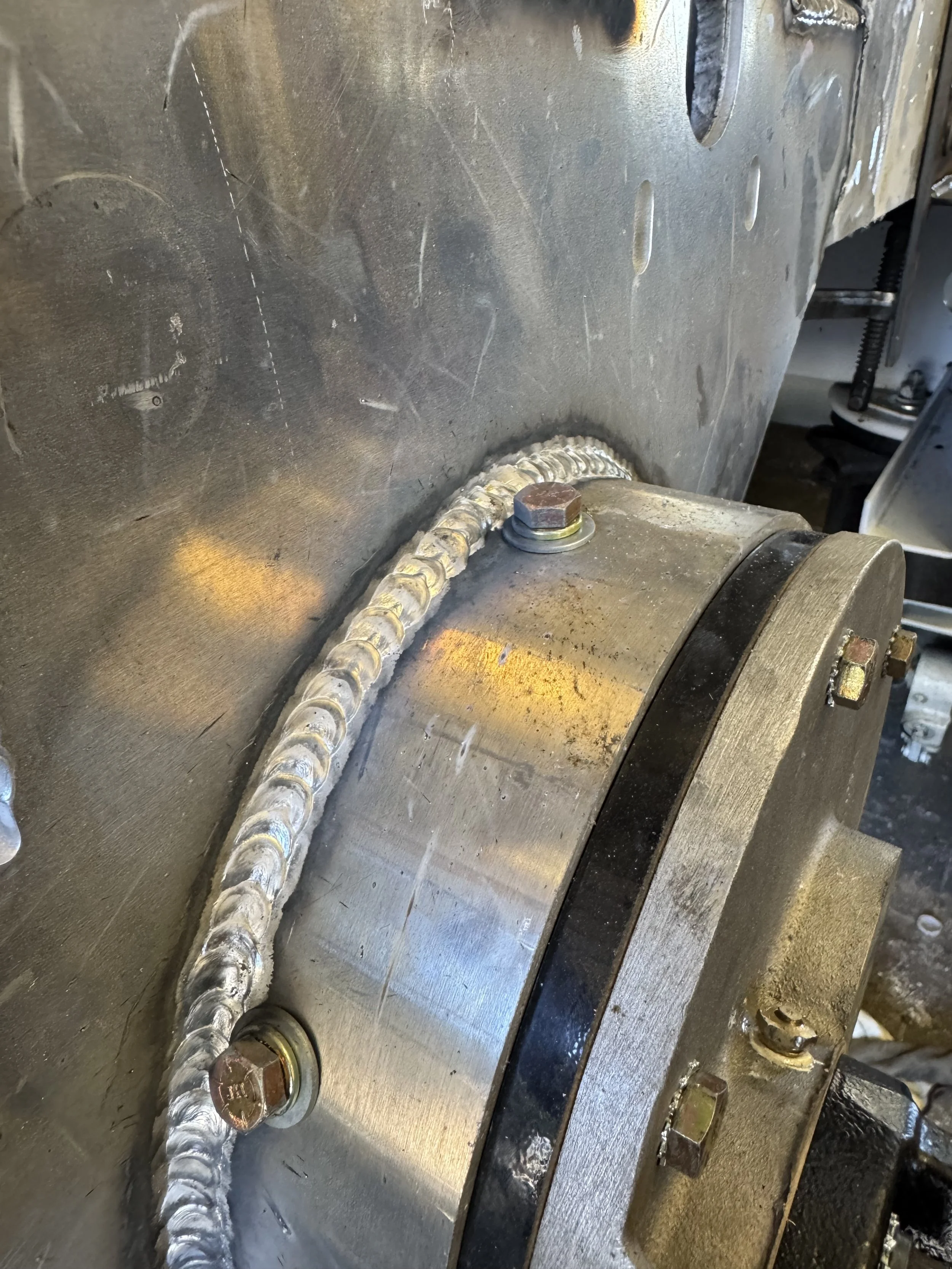 Close-up of a welded metal pipe with visible welding beads, attached to a larger metal component with bolts.
