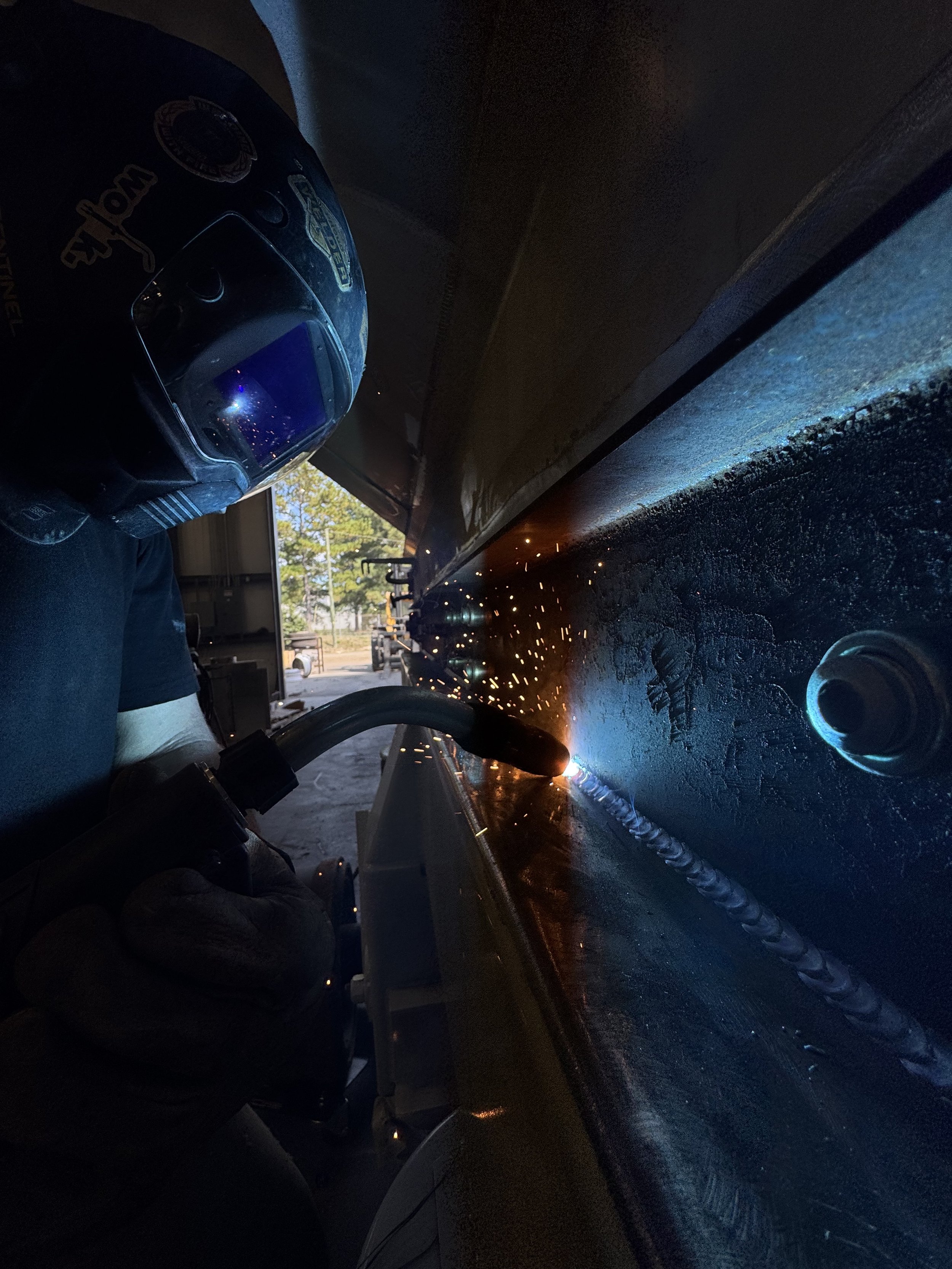 A person welding a metal structure outdoors at night, wearing a welding helmet and gloves, with sparks flying from the welding torch.