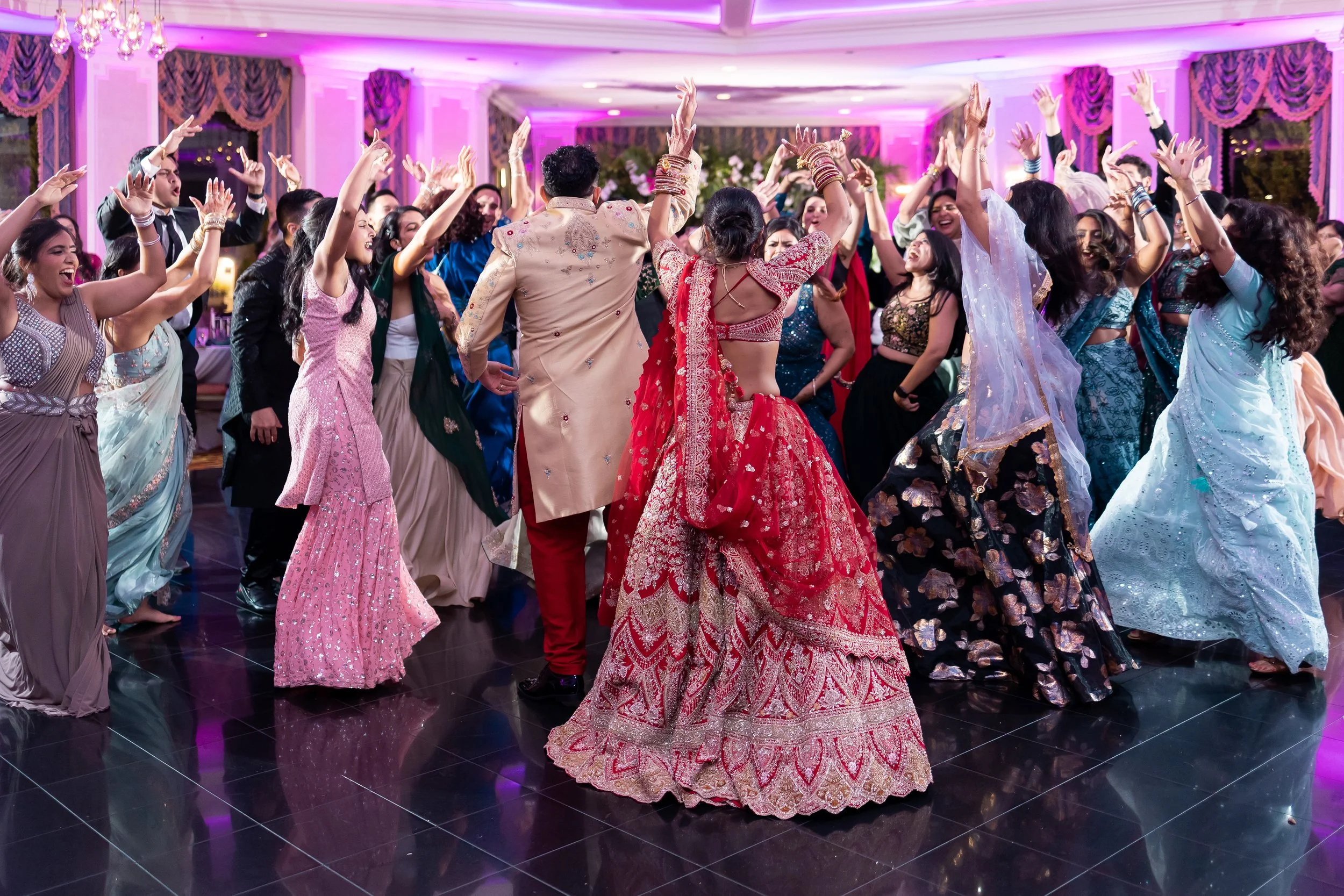 A group of people dancing together at a celebration, wearing colorful traditional Indian attire, in a decorated banquet hall with purple lighting.