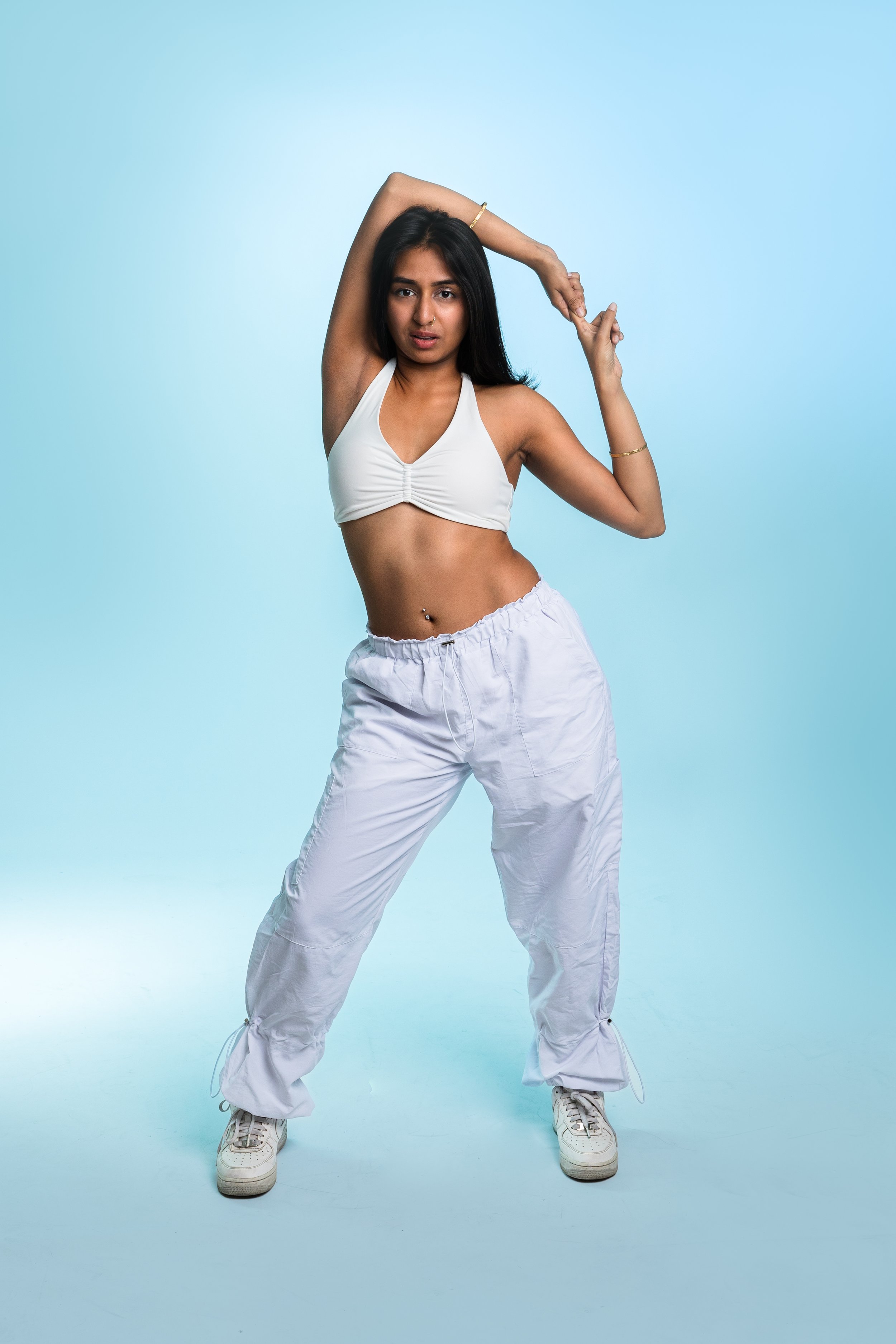 A young woman with long dark hair wearing a white sports bra, white baggy pants, and sneakers, striking a dance pose against a light blue background.