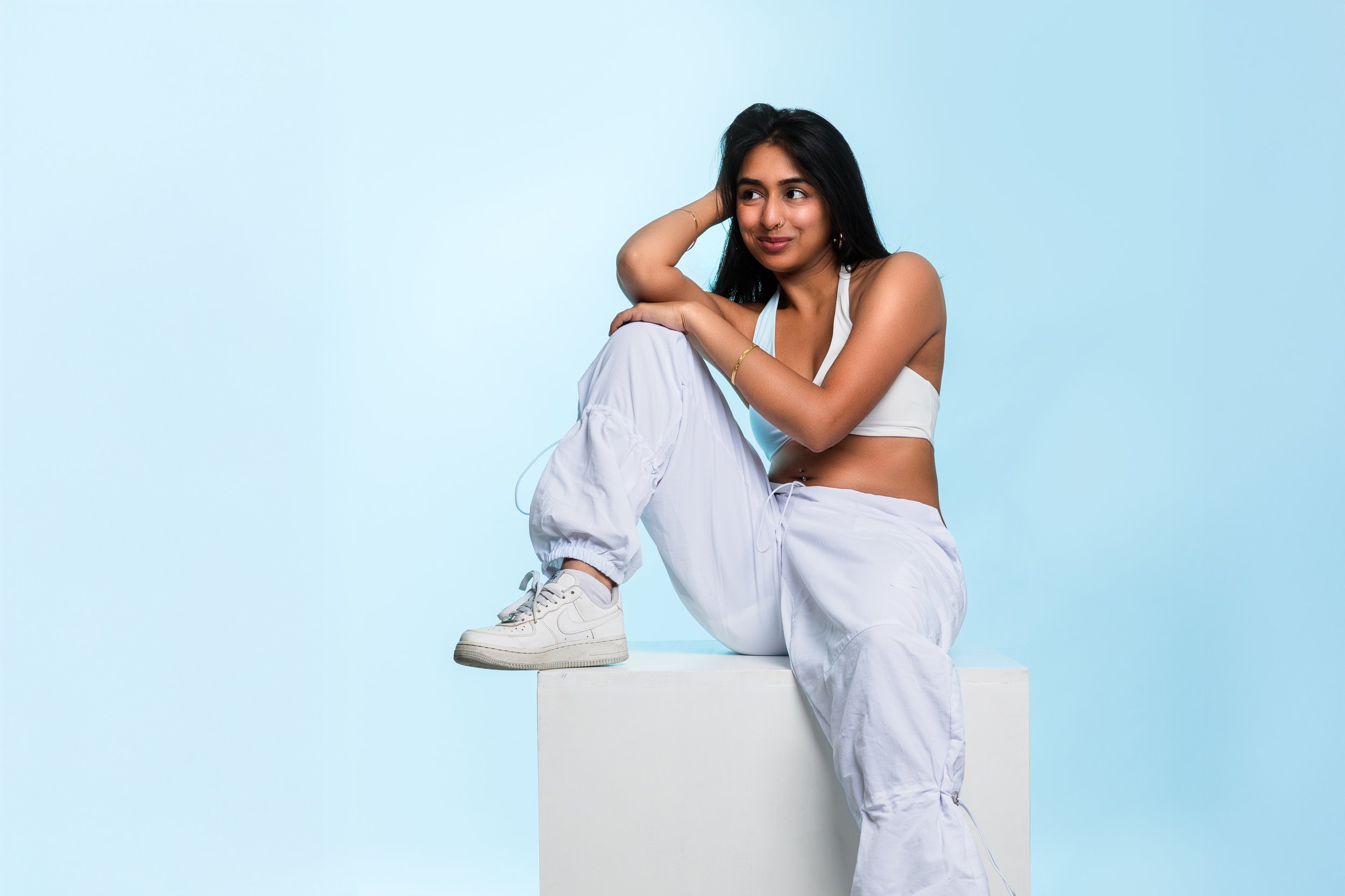A woman with long dark hair sitting on a white block against a light blue background, wearing a white crop top, white baggy pants, and white sneakers, looking to the side with a slight smile.