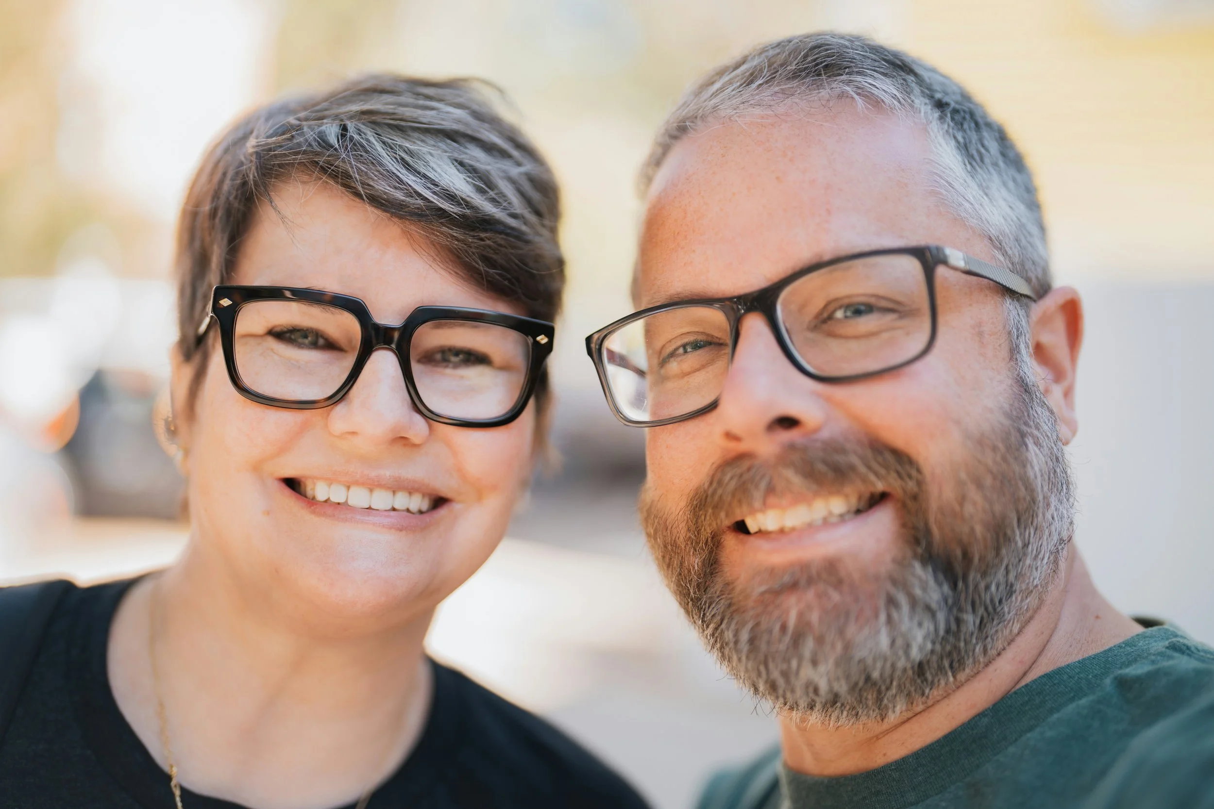 Close-up of a smiling woman and man wearing glasses, taking a selfie outdoors with a blurred background of trees and sunlight.