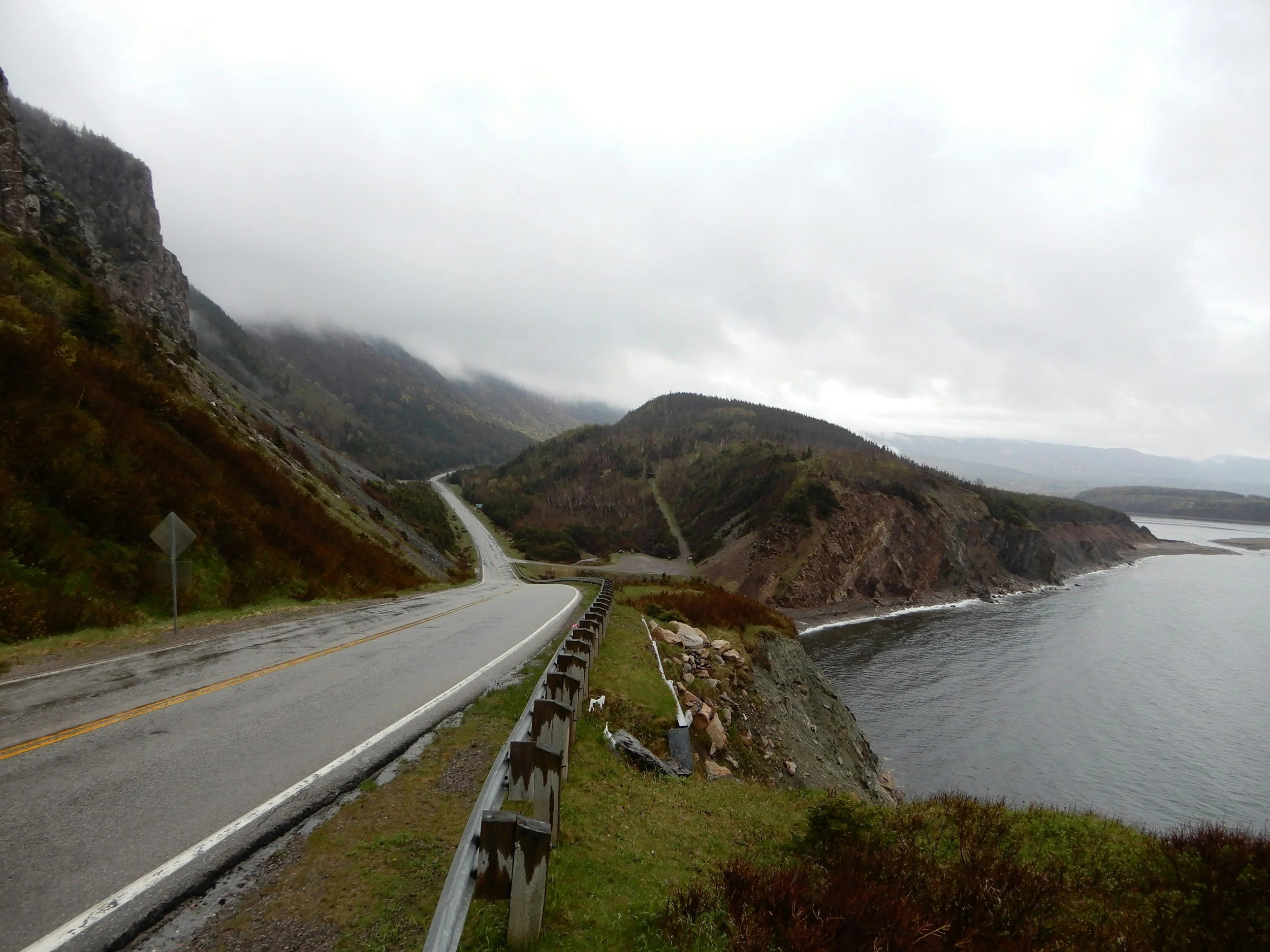 A winding coastal road runs alongside cliffs and the ocean on a cloudy day with fog hovering over the mountains.