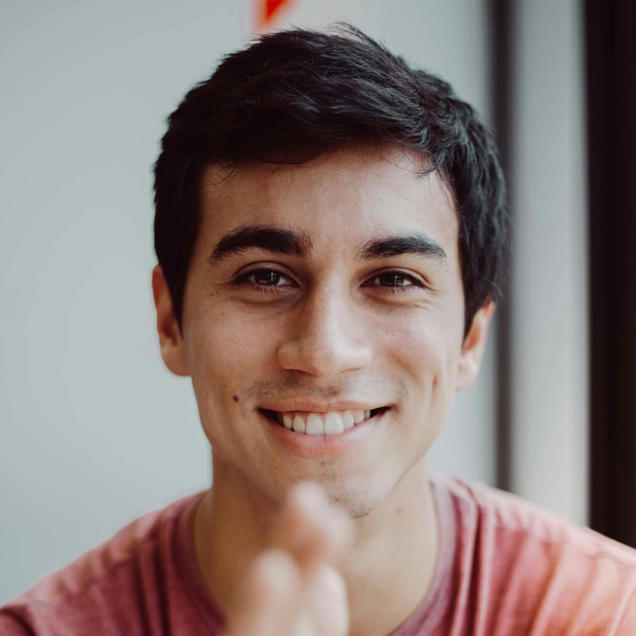 Close-up of smiling young man with dark hair and brown eyes, wearing a pink shirt.