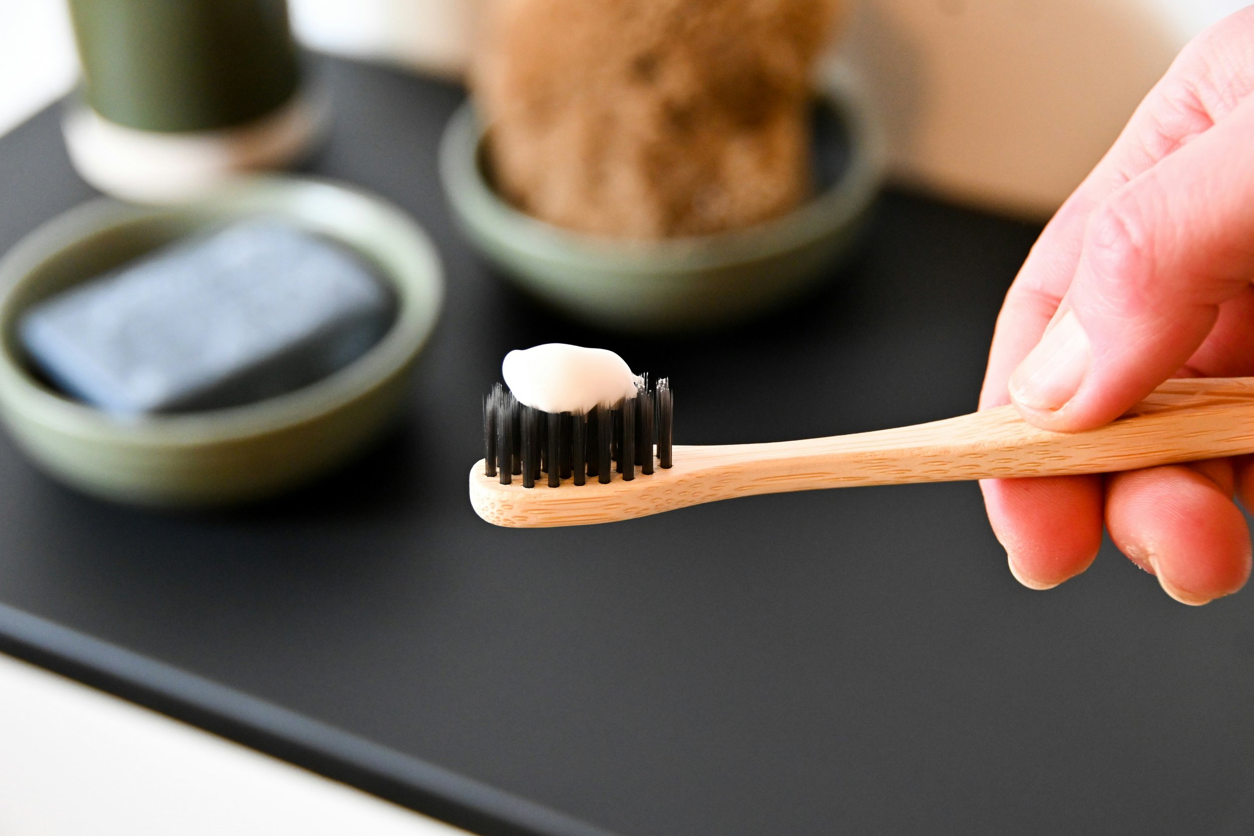 Close-up of a hand holding a wooden toothbrush with a dollop of white toothpaste on the black bristles, with small containers of black, green, and brown substances in the background.