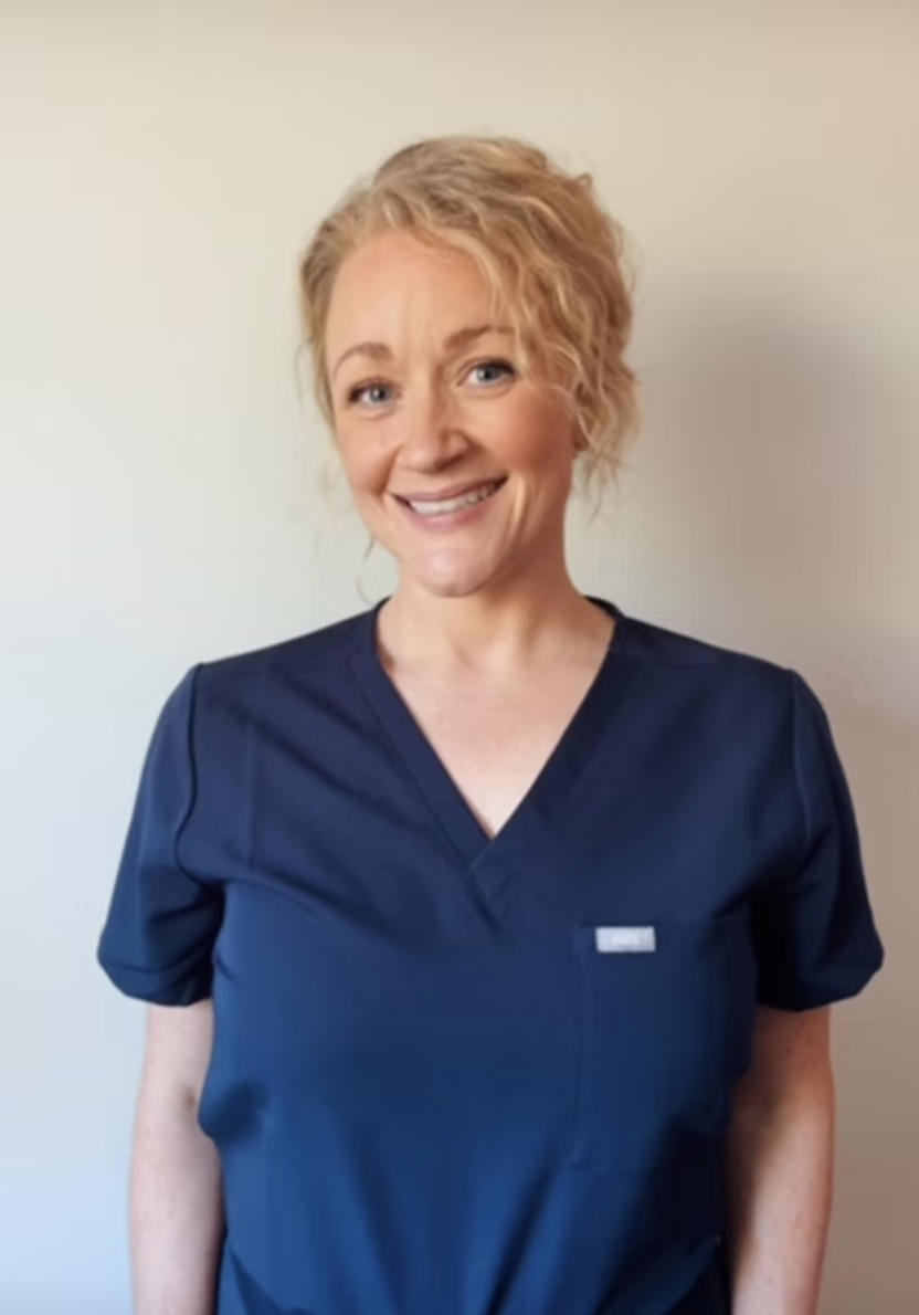 A woman with short, curly blonde hair wearing navy medical scrubs, smiling at the camera against a plain light background.